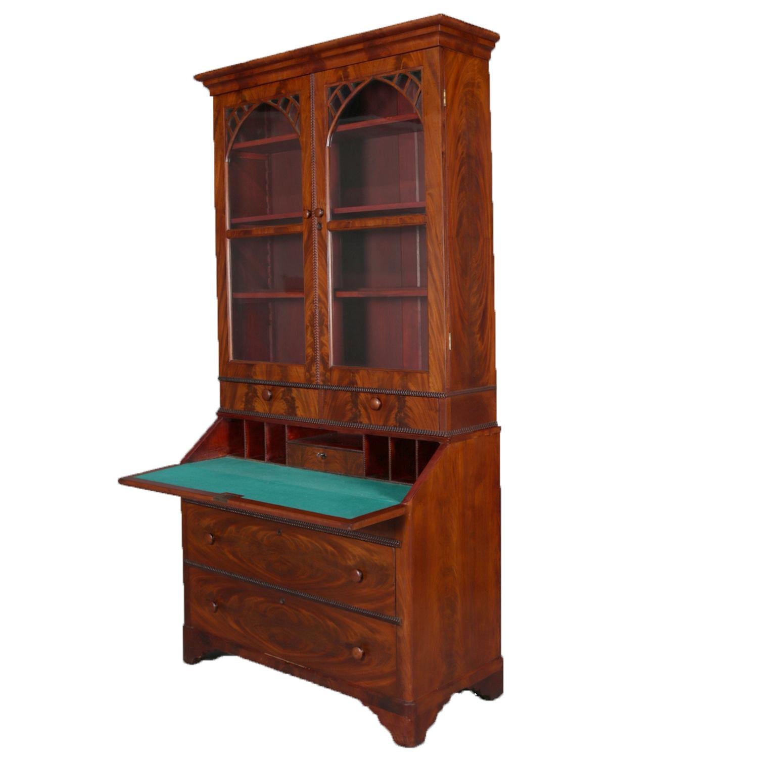Antique American Empire flame mahogany secretary features upper bookcase with double fretwork and arch form glass doors opening to shelved interior and over secretary with drop front desk having felt writing surface and pigeon holes over case with