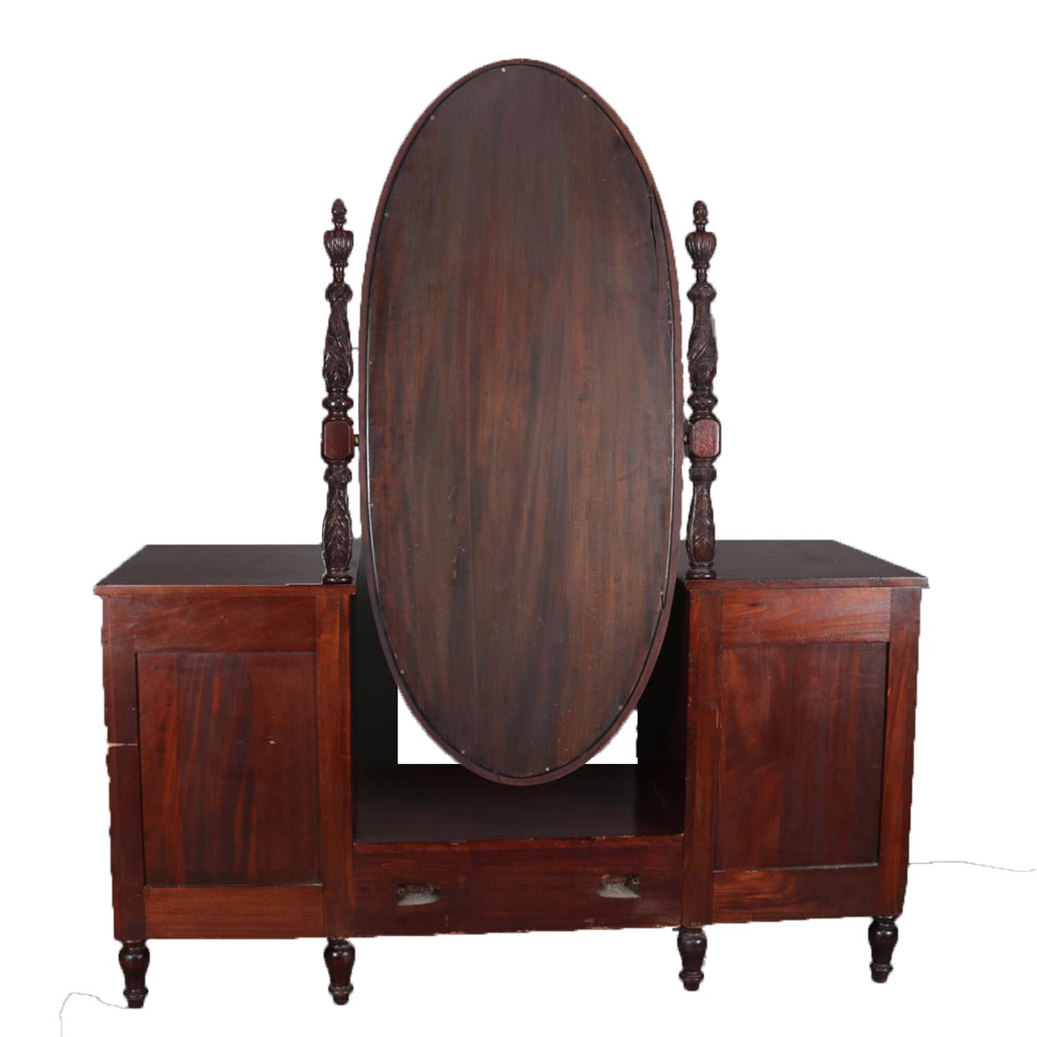 Antique American Empire dressing table feature mahogany construction with central elongated oval mirror flanked by deeply carved supports having central floral and acanthus, over central platform flanked by drawer columns each having an upper convex