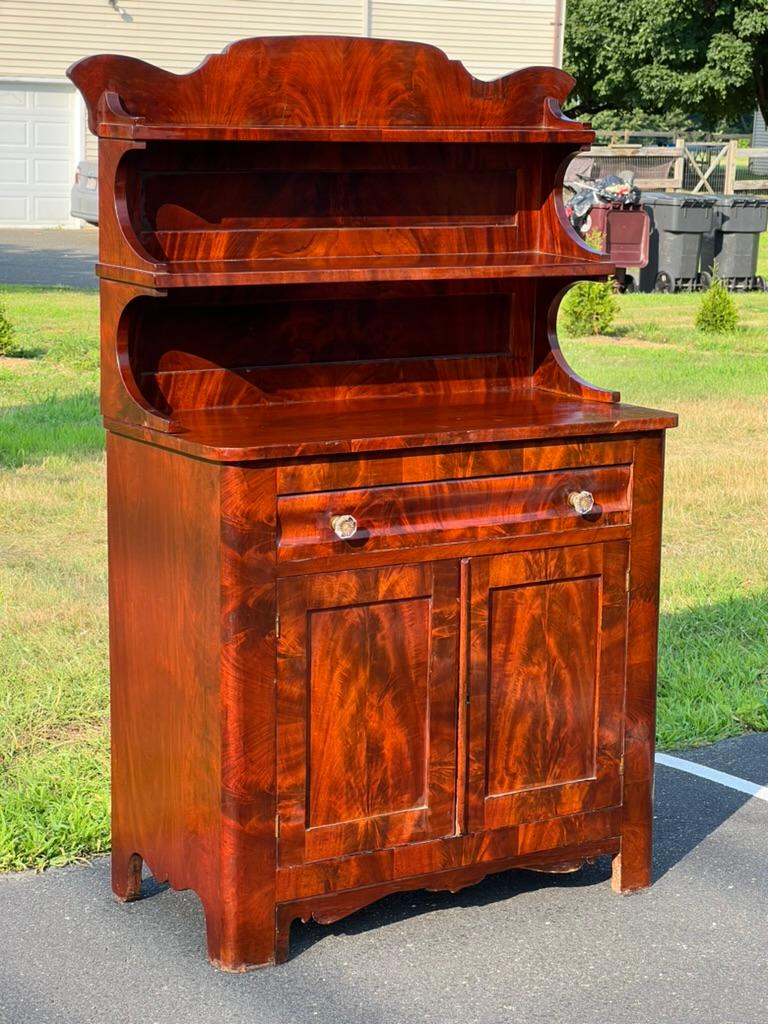 Immerse yourself in the refined elegance of the early 19th century with this Antique American Empire Flame Mahogany Diminutive Cupboard. This exquisite piece showcases the flame mahogany's striking grain pattern, characterized by its vibrant and