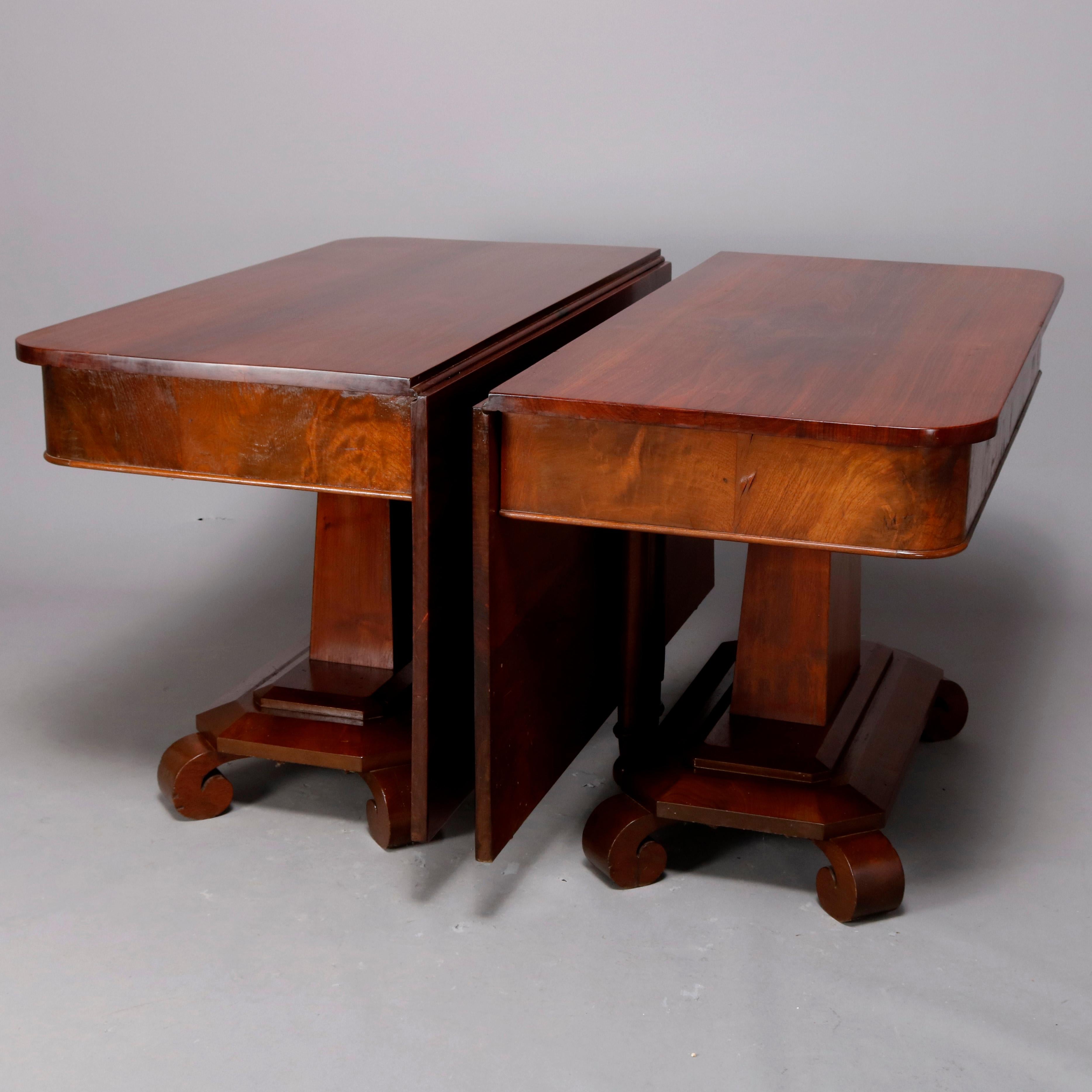 An antique American Empire flame mahogany banquet table offers divided form, each with deep skirt, flared pedestal and raised on stepped base with scroll form feet, drop leaves raise and are supported by turned and folding leg, circa