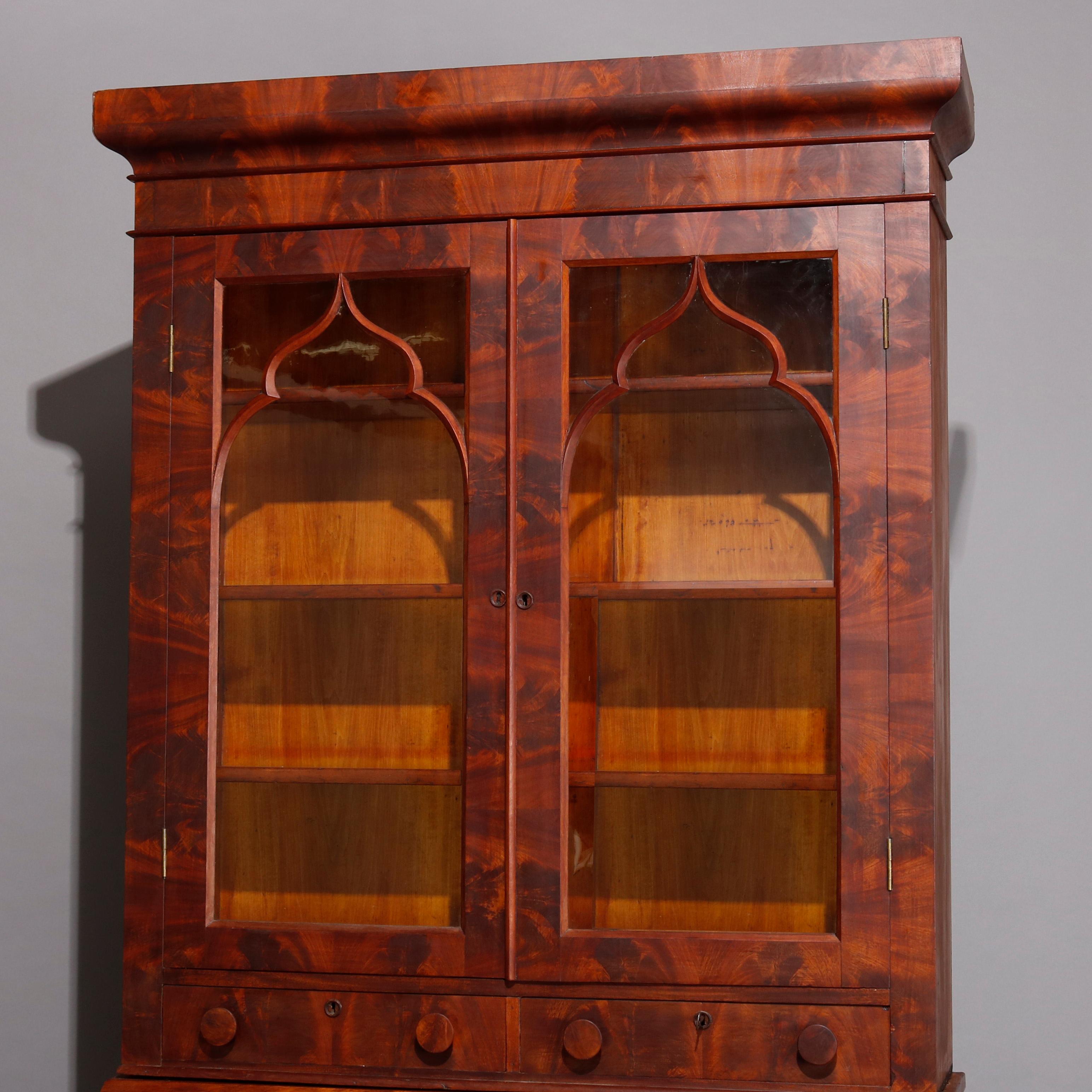 An antique American Empire secretary offers deeply striated flame mahogany construction with upper bookcase having double glass doors with arch form filigree and two drawers surmounting lower case with drop front desk over ogee frieze drawer and two