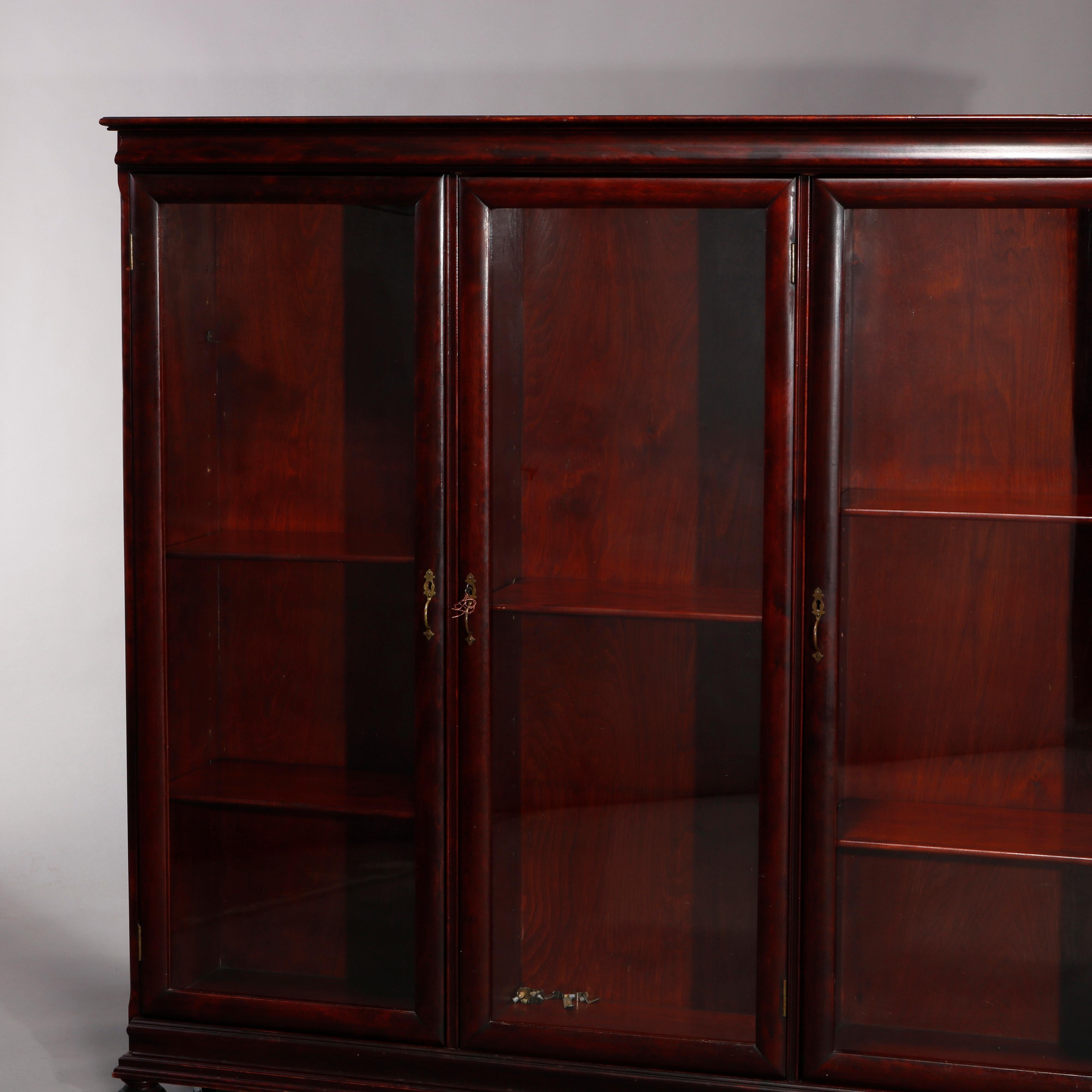 An antique American empire triple bookcase offers flame mahogany construction with ogee frieze surmounting case with three glass doors opening to divided and shelved interiors, raised on bun feet and having cast bronze hardware, locking and with