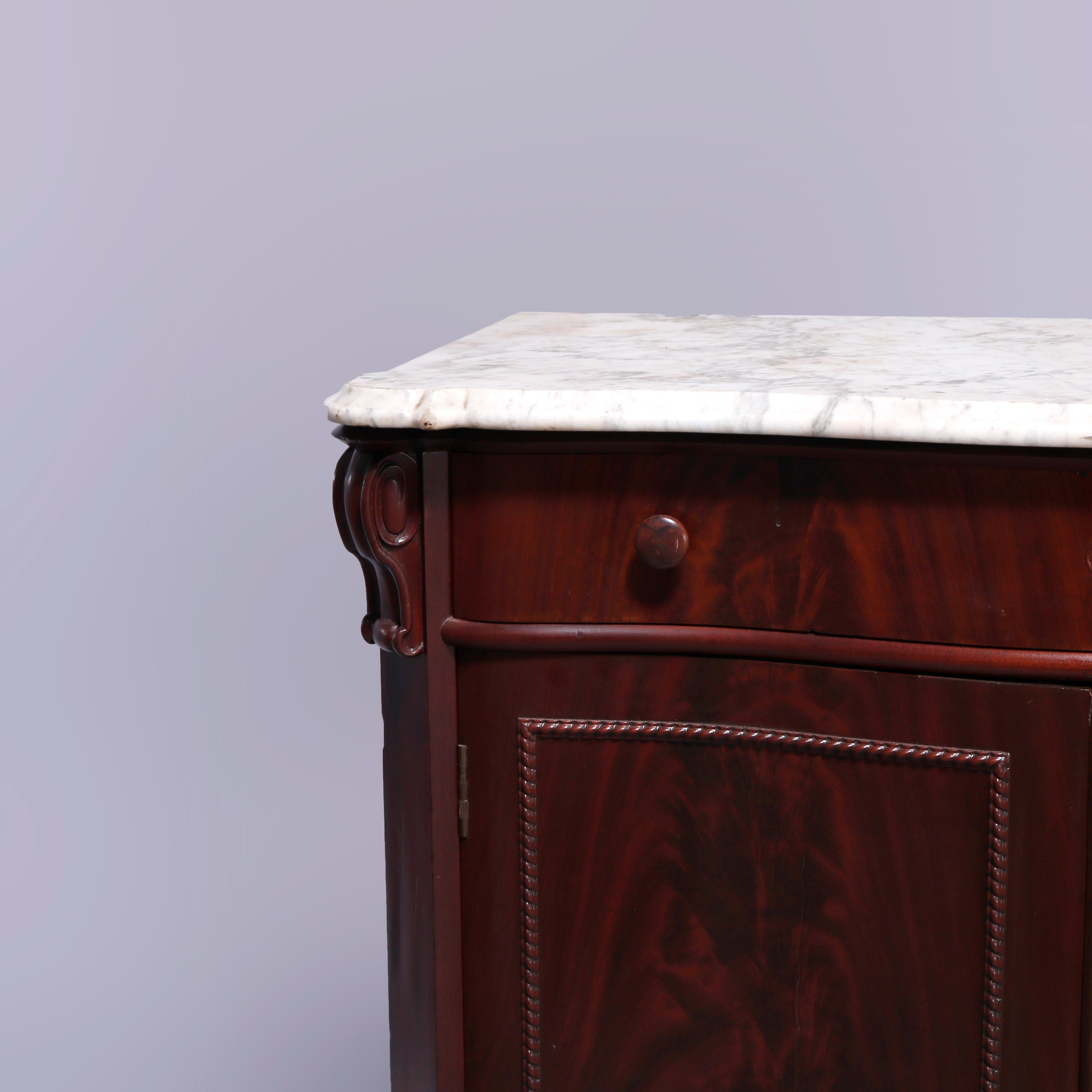 An antique American Empire commode offers shaped and beveled marble top surmounting flame mahogany in serpentine form with frieze drawer flanked by caved scroll form corbels and over lower double door cabinet with bead trim and opening to shelved