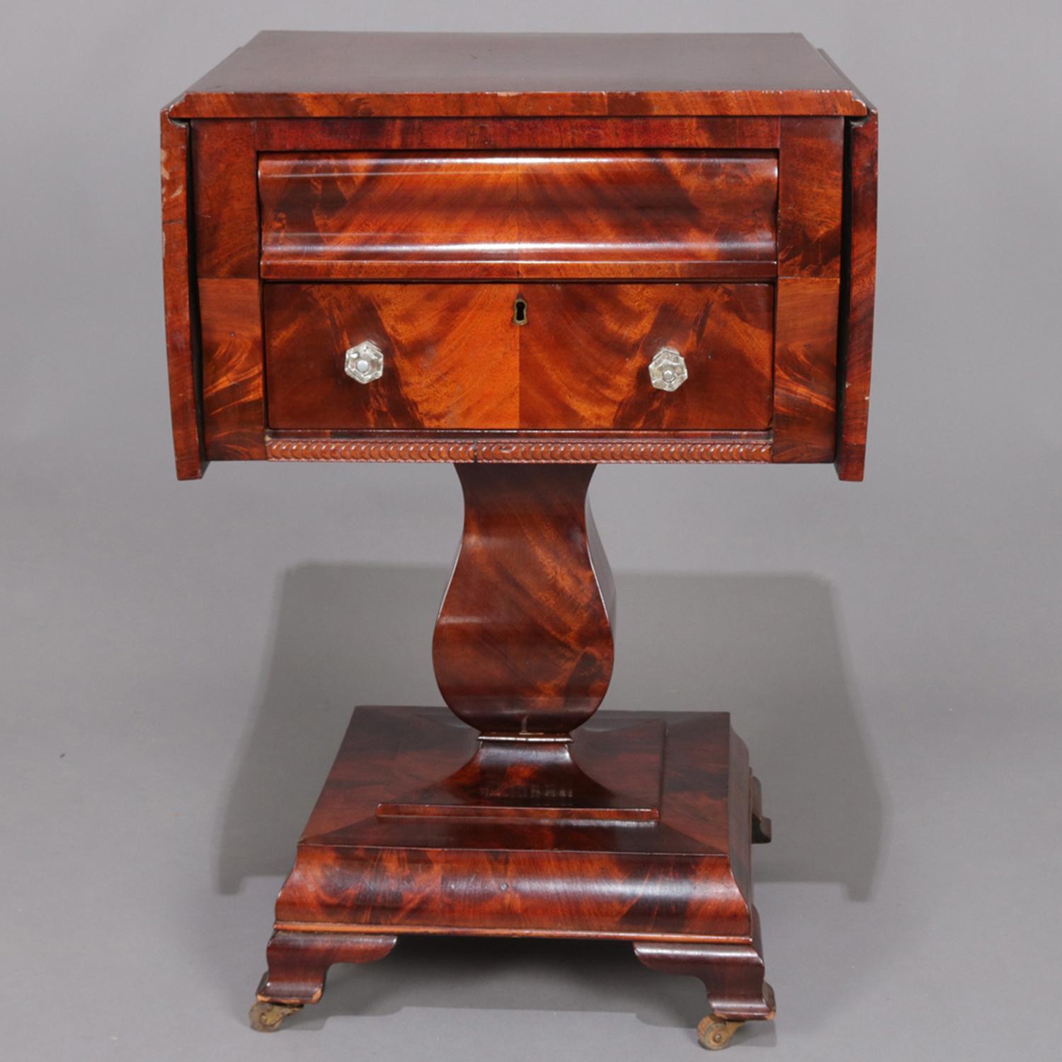 Antique American Empire sewing stand features flame mahogany construction with bookmatched two-drawer case and drop-leaf top seated on urn form pedestal above stepped and footed base, circa 1840

Measures: 30