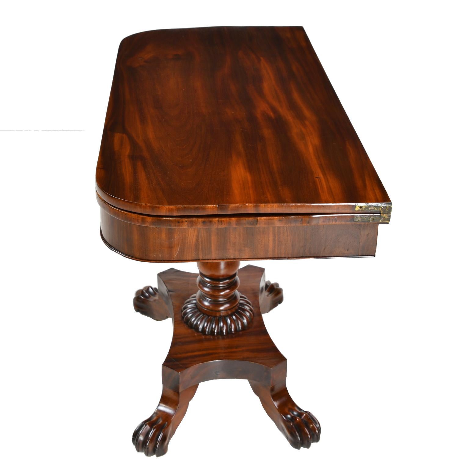 A beautiful American Empire card or games table in West Indies mahogany with hinged top opening to a square over a rectangular base with turned column that rests on a quatre-form base on carved lions paw feet. A cartouche with carved foliage