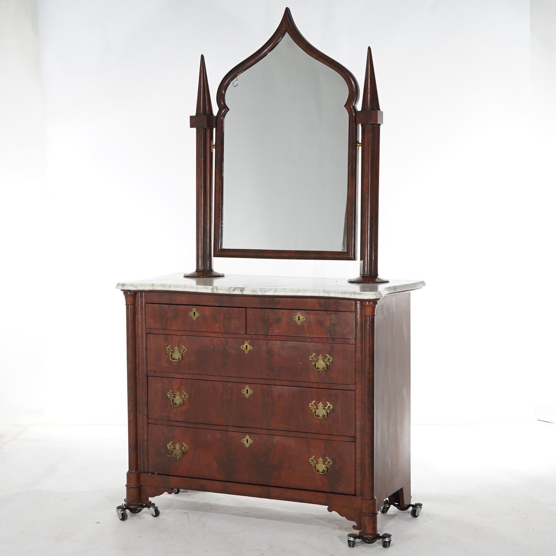 An antique American Empire Gothic Revival chest of drawers offers flame mahogany construction with cathedral arch form mirror with flanking spire supports over shaped marble top over serpentine chest having two smaller upper and three lower long