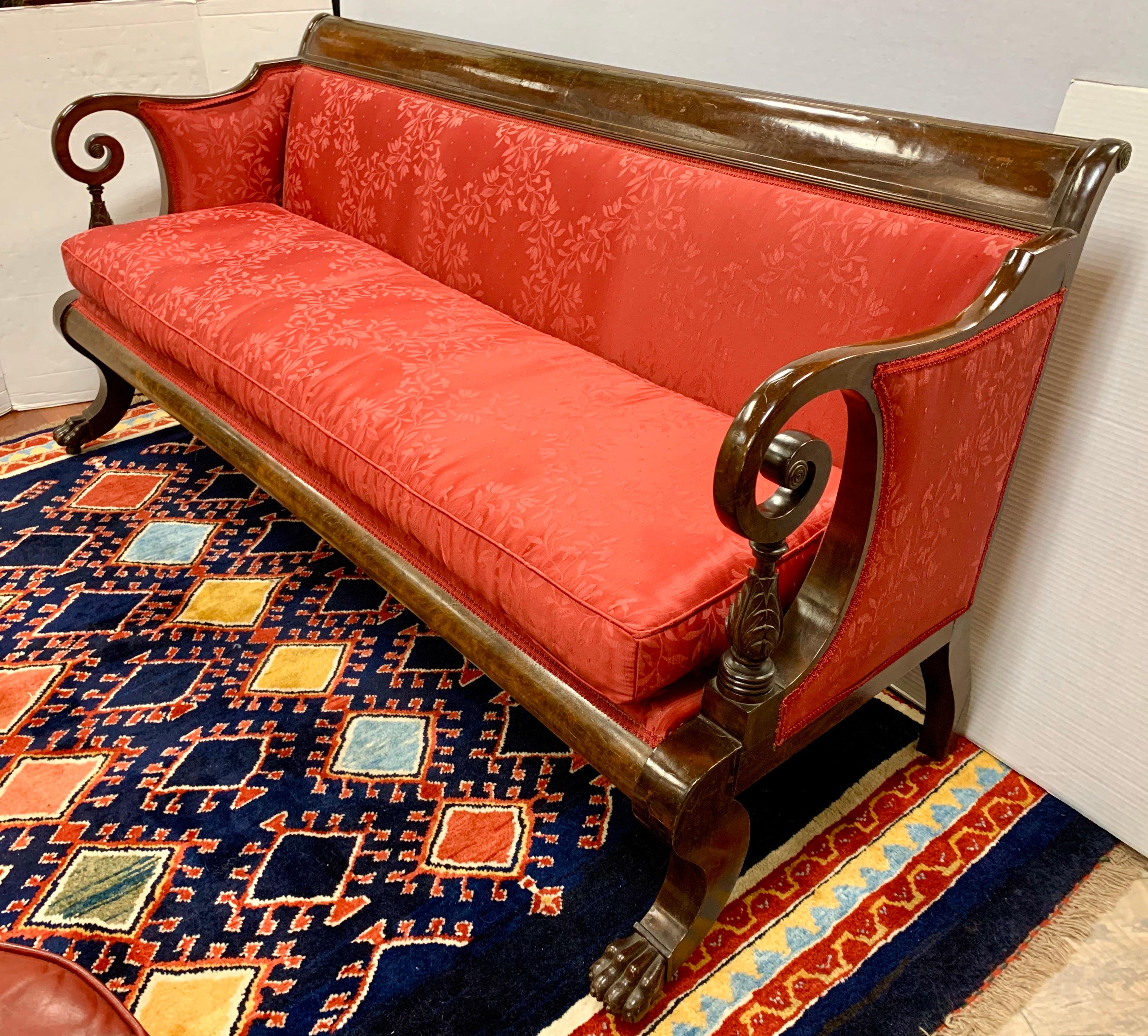 Antique 19th century empire sofa made of solid mahogany with scroll arms and carved lion paws with castors on front, upholstered in a red silk fabric. Down seat cushion makes it very comfortable.  Circa New York 1820-1935.