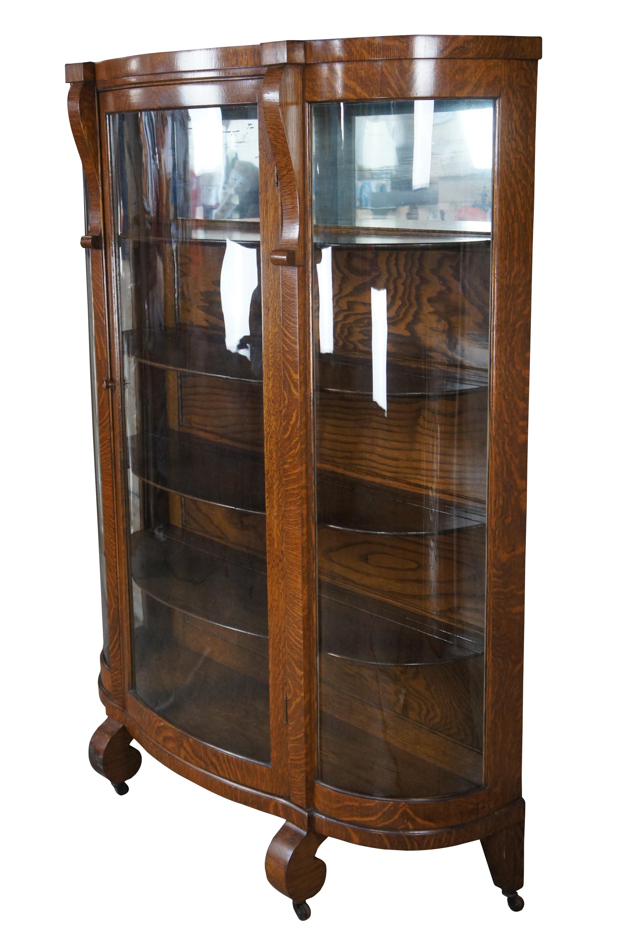 Antique Victorian Era American Empire demilune curved glass curio cabinet. Made from quartersawn oak with scrolled accents along the front leading to scrolled feet over casters.. Includes four shelves with plate groves and a mirror along the top.