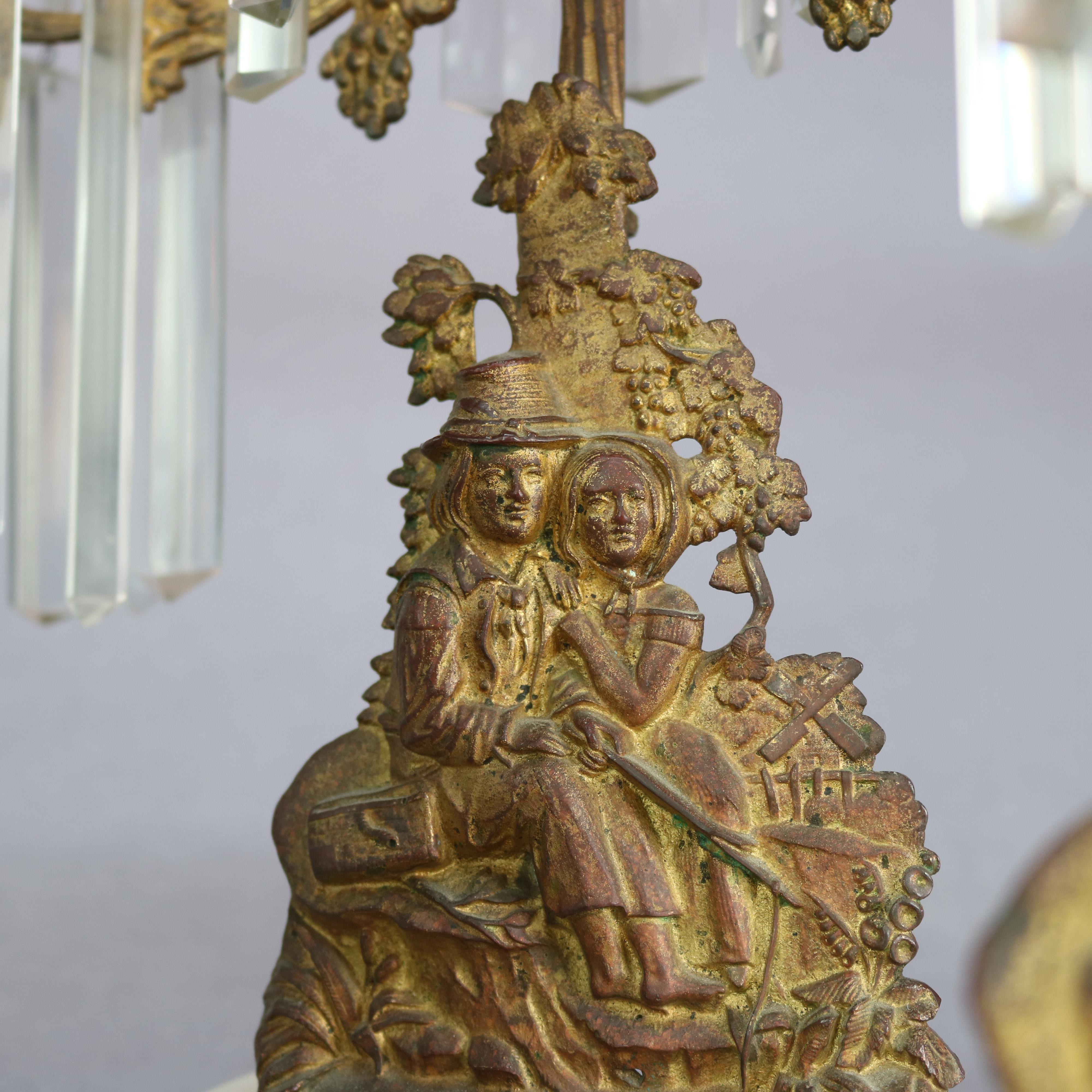 An antique figural American girandolle set offers central candelabra with flanking candlesticks each having cast brass base with courting scene in countryside setting, seated on marble bases and with hanging cut crystal bobeches highlights,