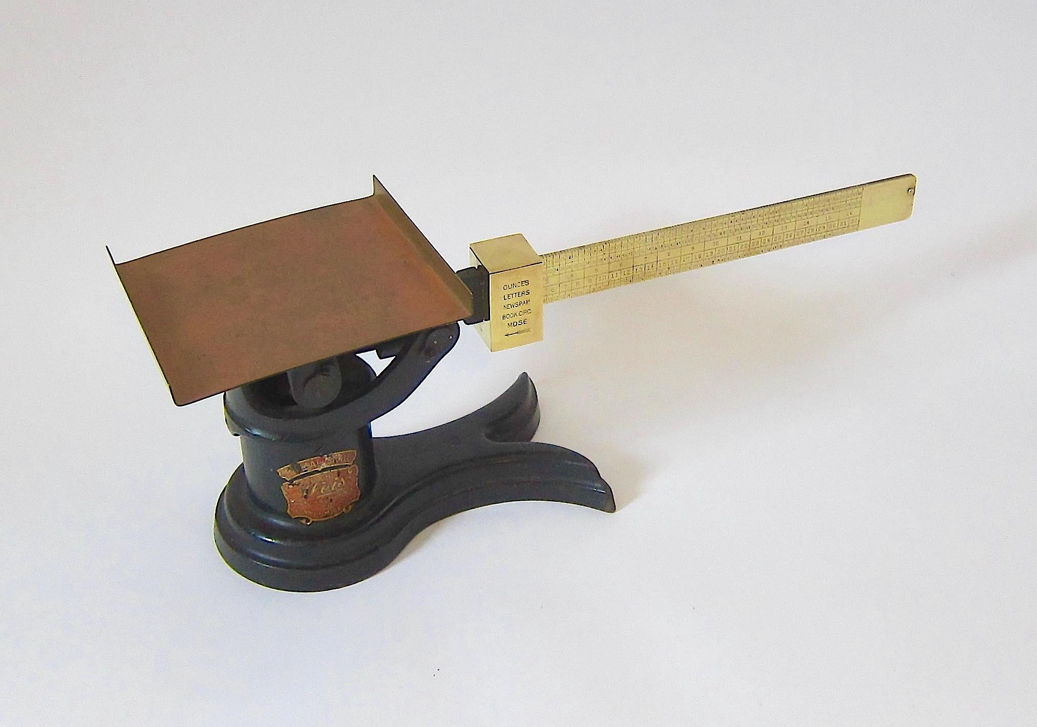 An antique American post office scale in working condition by Weis Manufacturing Company of Monroe, Michigan, USA. The base is shaped like a fishtail in heavy cast iron with a tray, arm, and counter weight of cast brass. The strong silhouette and