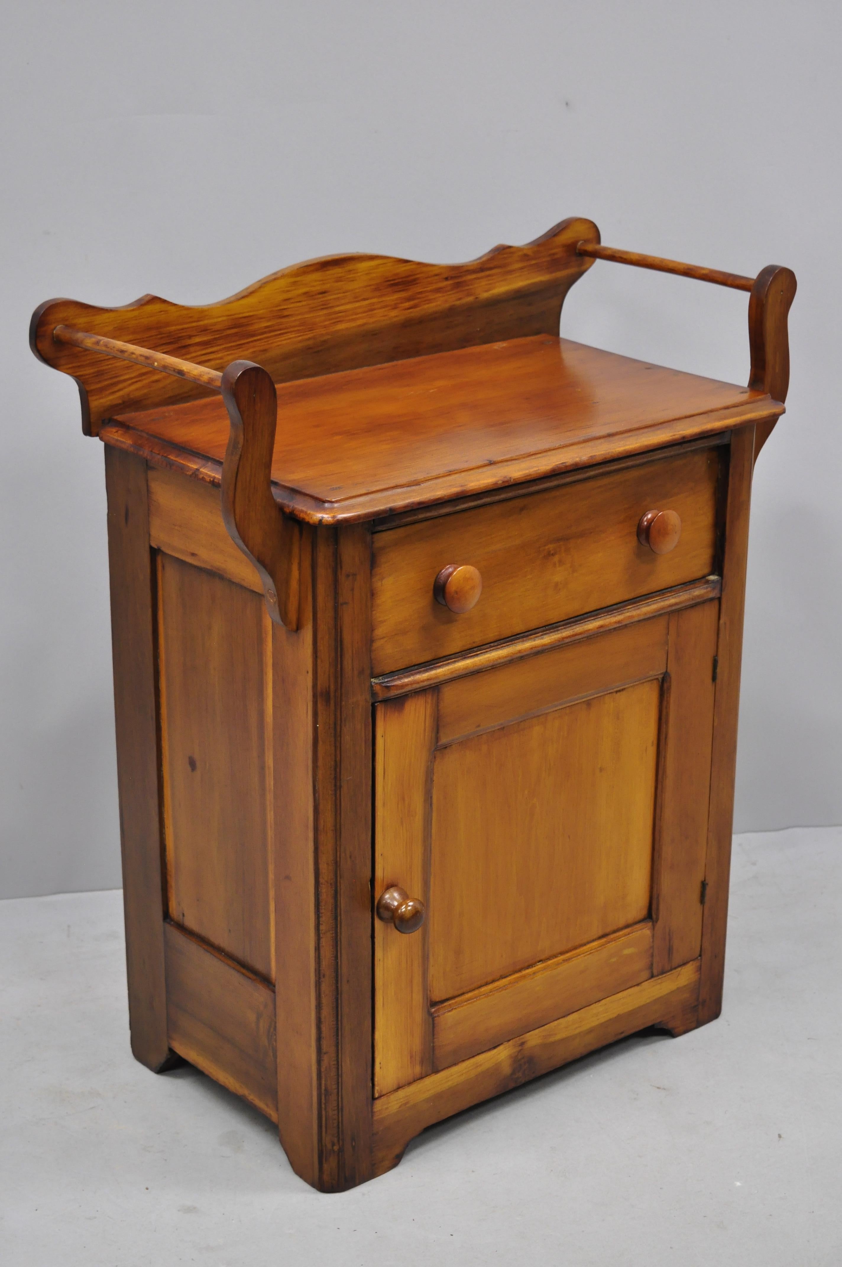Antique American primitive 19th century pinewood washstand cabinet end table. Item features shaped backsplash, 2 towel bars, solid wood construction, beautiful wood grain, 1 swing door, 1 dovetailed drawer, very nice antique item, quality American