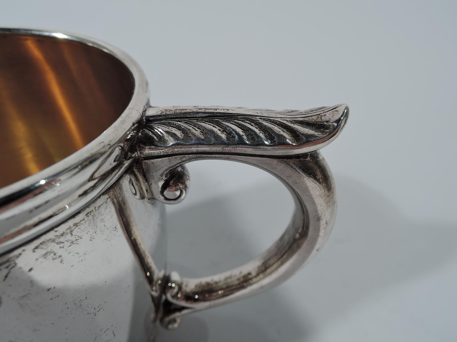 Sterling silver christening mug. Made by Durgin (later part of Gorham) in Concord, New Hampshire, circa 1900. Barrel form with molded rim and leaf-capped scrolled handle. At base tooled Classical ornament with alternating scallop shells and