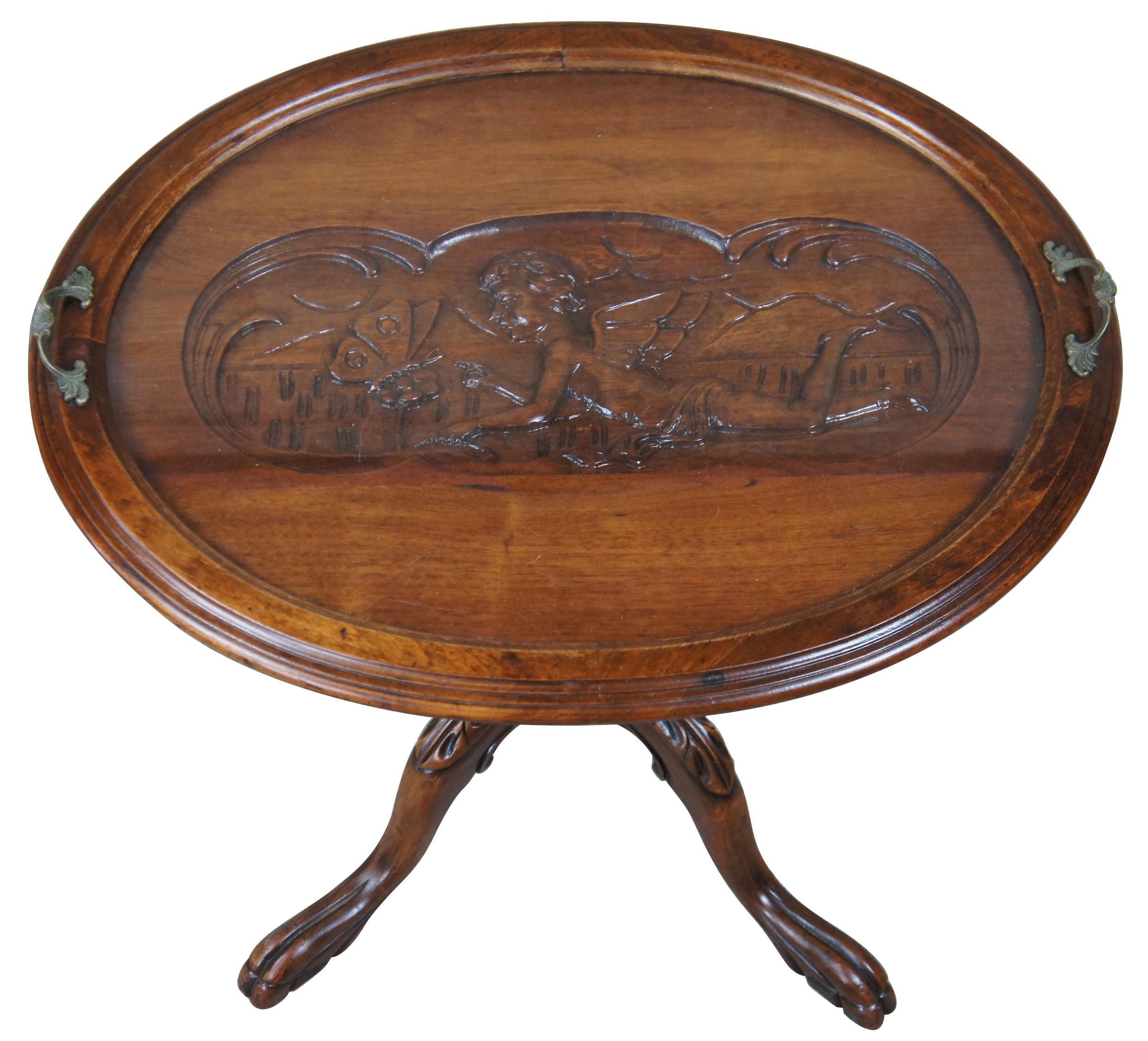 Antique late Victorian carved walnut pedestal tray table. Features a central figural scene with a boy angel and butterfly. The table sits atop four paw feet. The tray features glass center and brass handles, making it nice for serving.
  