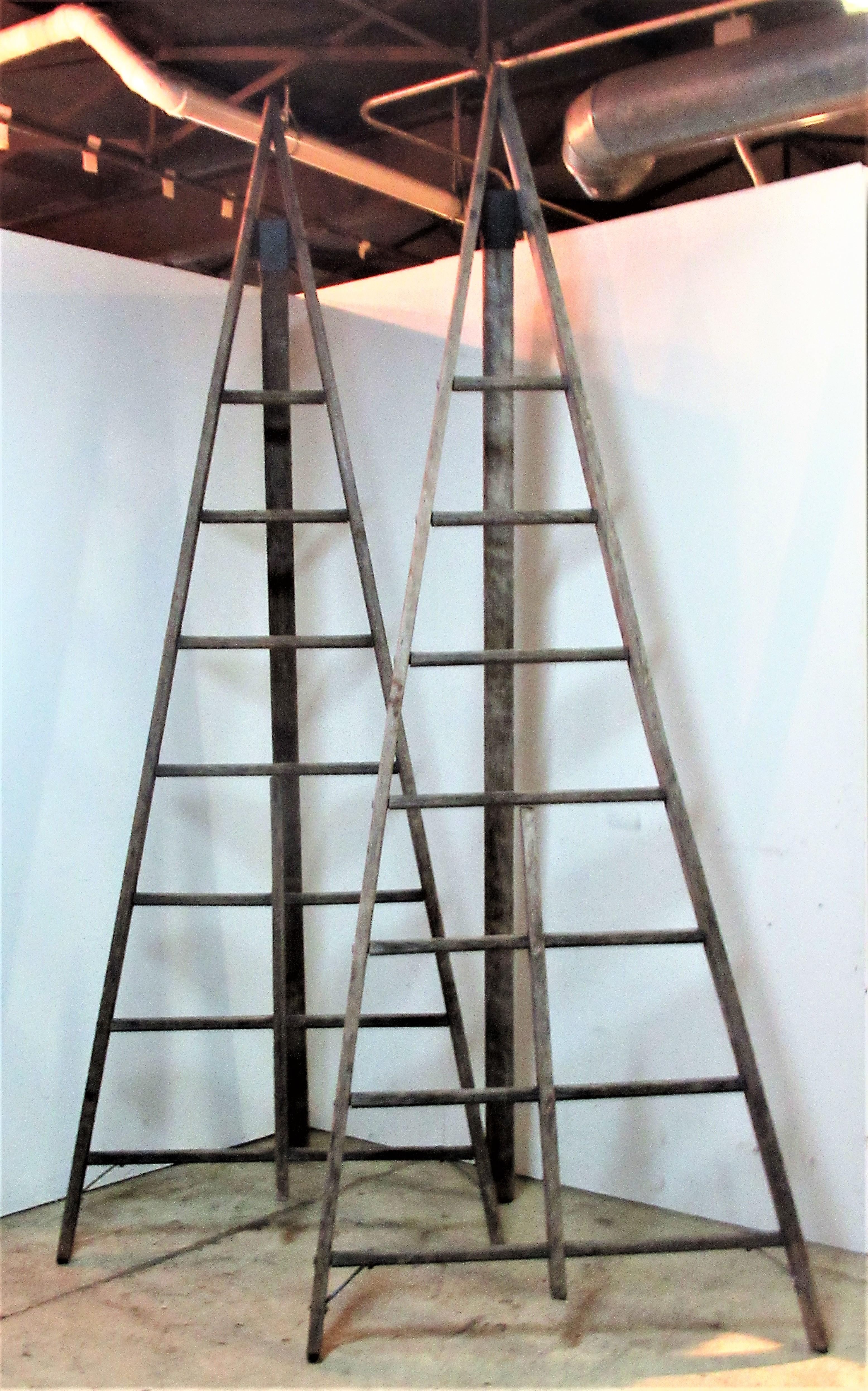 A-frame form folding peak top wood and riveted steel industrial orchard harvest work ladders with beautifully aged old weathered driftwood gray surface and great architectonic sculptural form. American, circa 1940-1950. Ladders measure folded 110