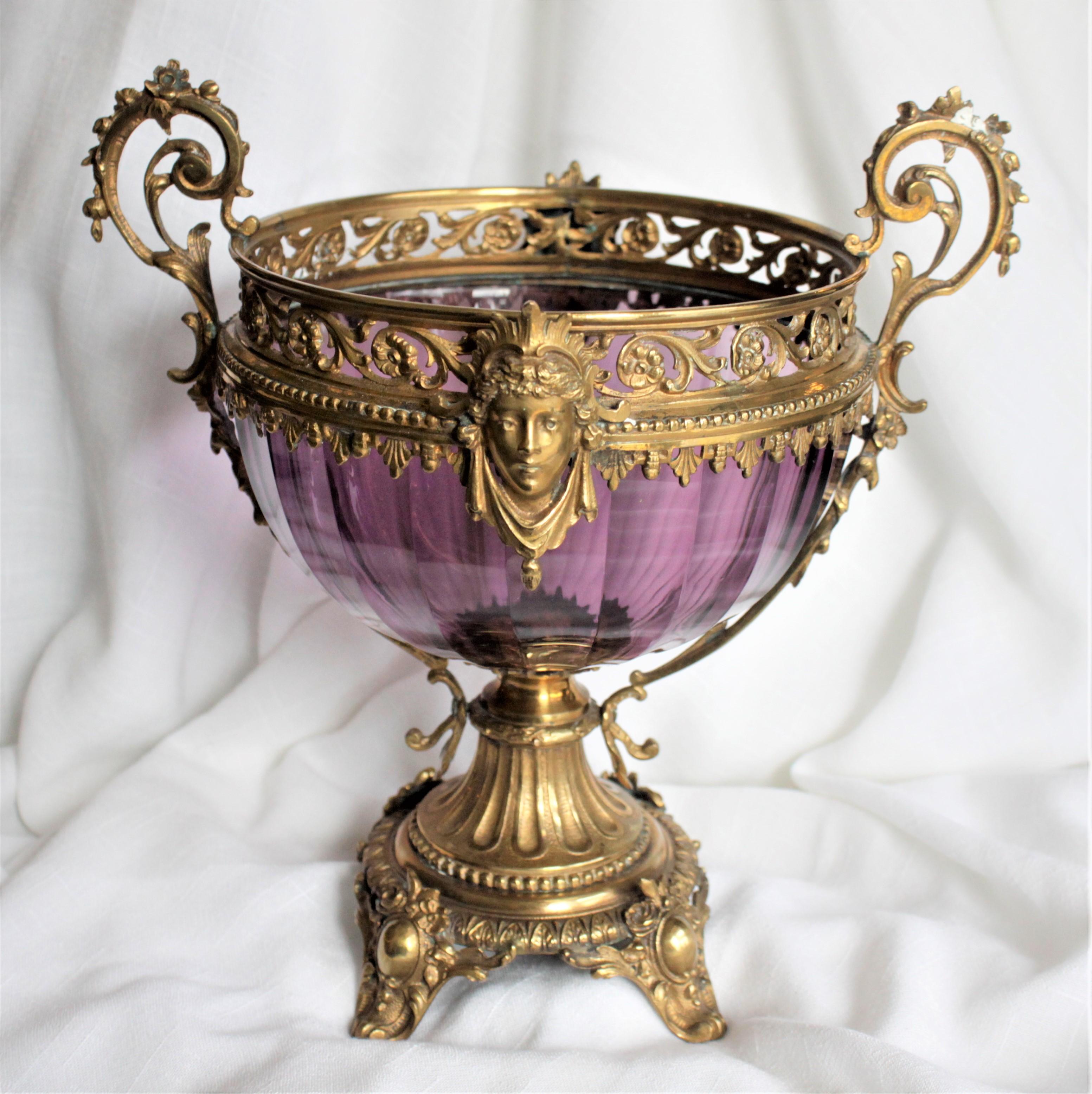 This antique amethyst glass pedestal vase is unmarked but presumed to have been made in England in circa 1880 in the Victorian style. This compote features very elaborate and ornate gilt bronze mounts with large scrolled handles and a footed base.