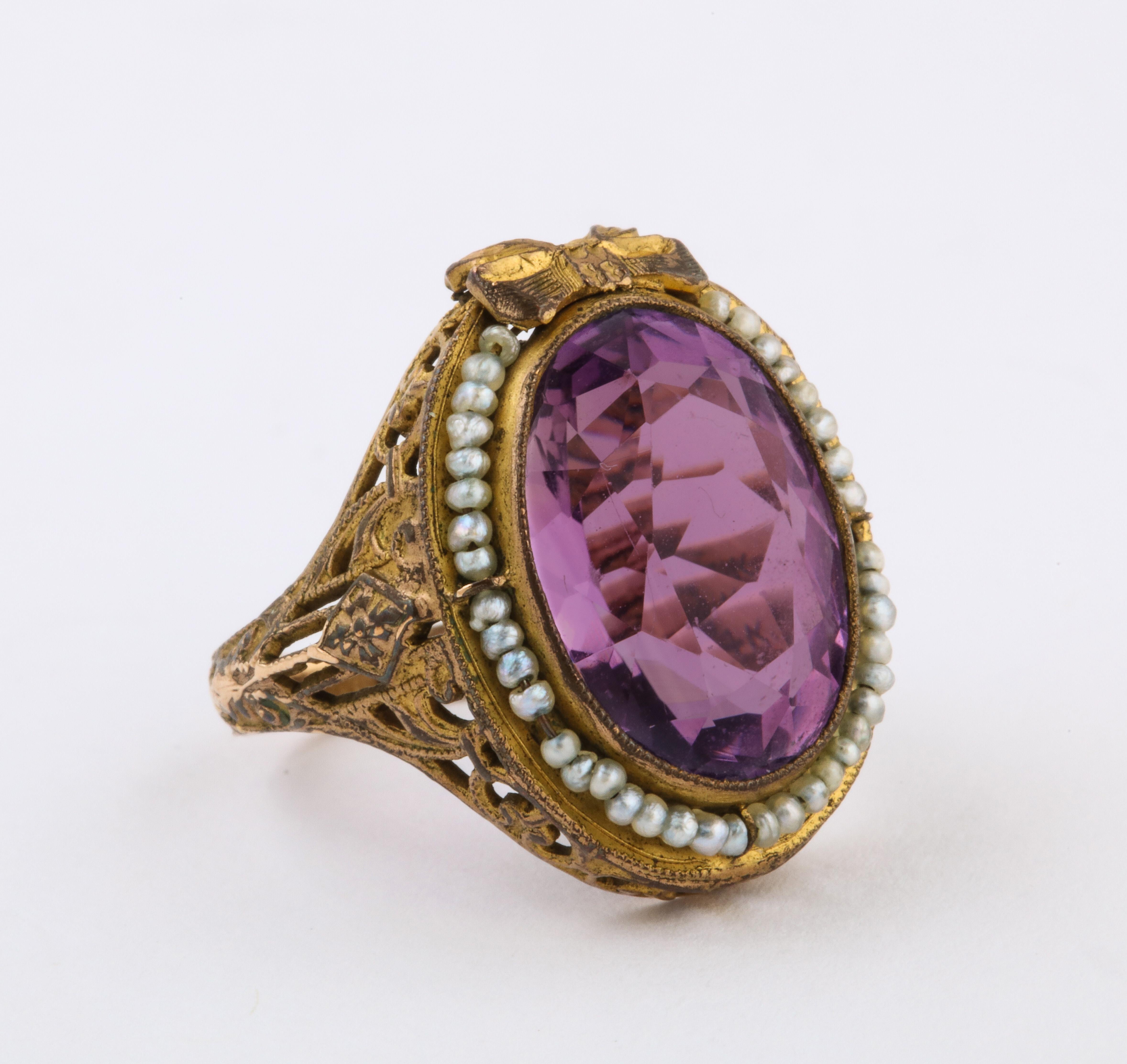 Of regal design set with an oval-cut amethyst of light purple color weighing approximately 6 carats, capped by a bow and surrounded by a border of seed pearls, mounted in an intricate openwork brass and gold setting. 

size  4. Amethyst measures