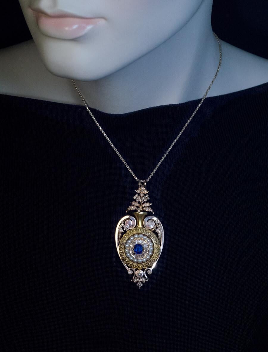 Circa 1870

This mid Victorian era large locket pendant is modeled as a stylized amphora-shaped vase with flowers. The pendant is finely crafted in yellow and rose 14K gold. It is centered with a cushion cut blue sapphire encircled by old mine cut