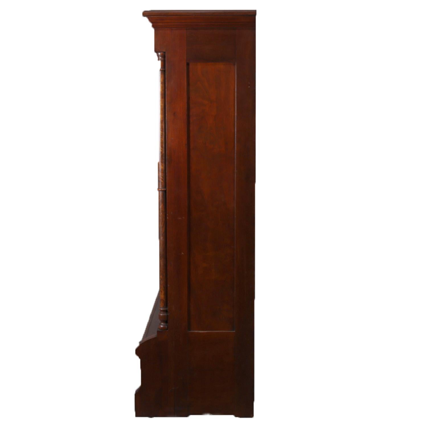 An antique and large Eastlake step-back bookcase features walnut construction with carved crest over case with three glass doors opening to adjustable shelved interior and flanked by Corinthian column supports surmounting three burl inset storage