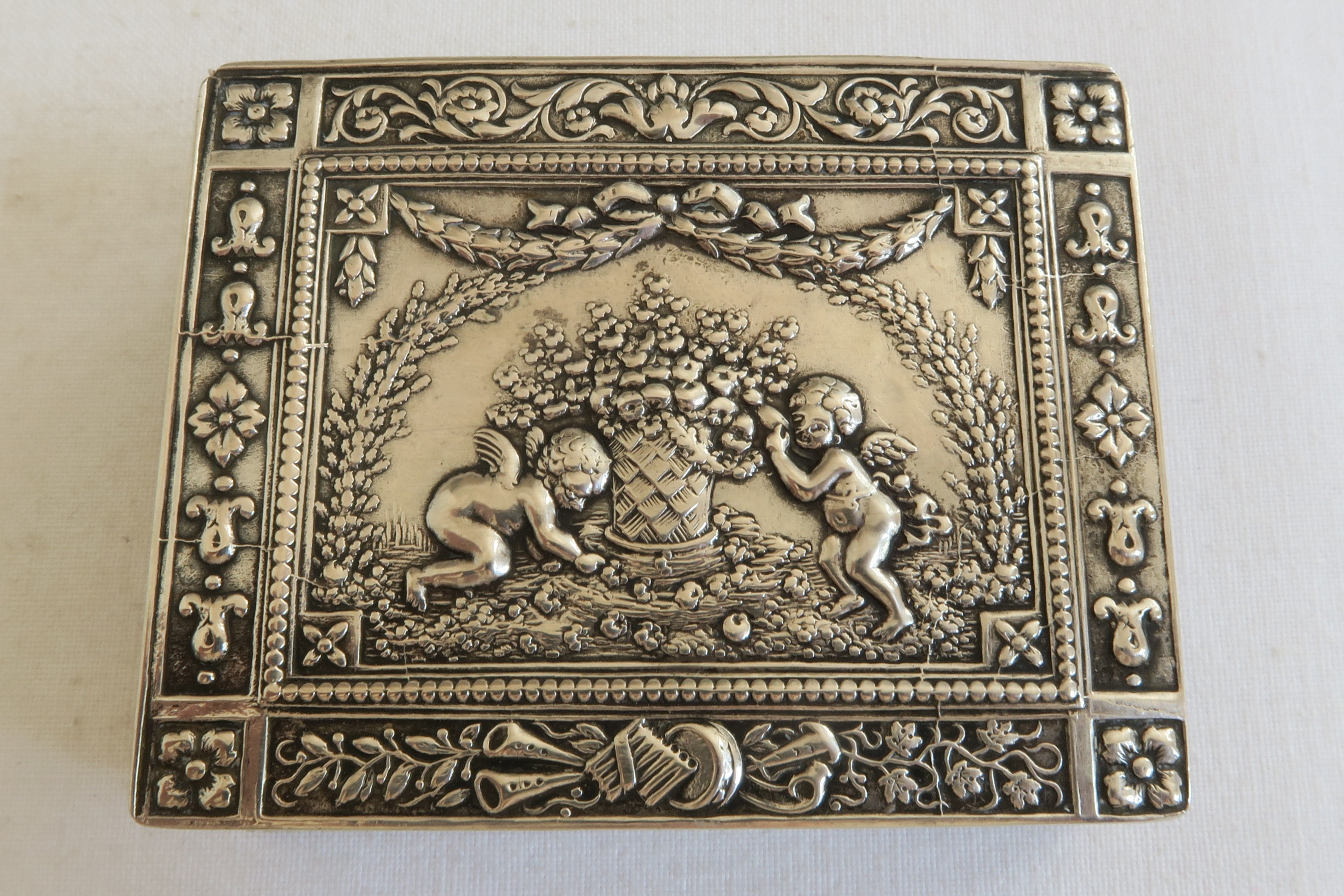 This beautiful snuff box is made from sterling silver. The lid and sides are covered in an intricate floral design with musical instruments and a scene of two playing cherubs in its center. The richly embellished box was manufactured in Germany and