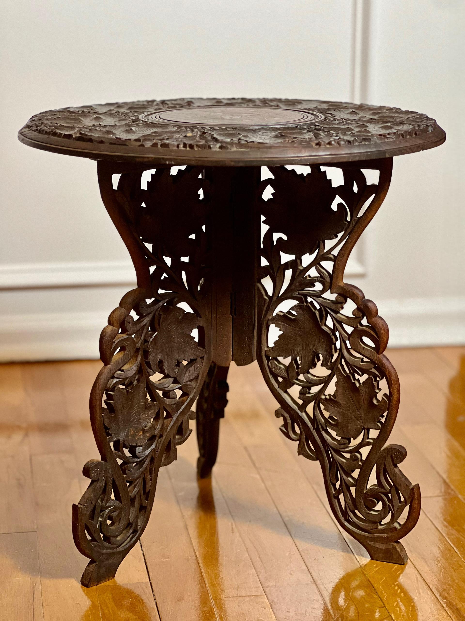 An early 20th century Indian foliate carved round tea table on a folding base. A tabletop with ornate relief carvings of grapevine leaves encircling a smooth center medallion with gold accents, rests upon a tripod form, hinged base with bold pierced