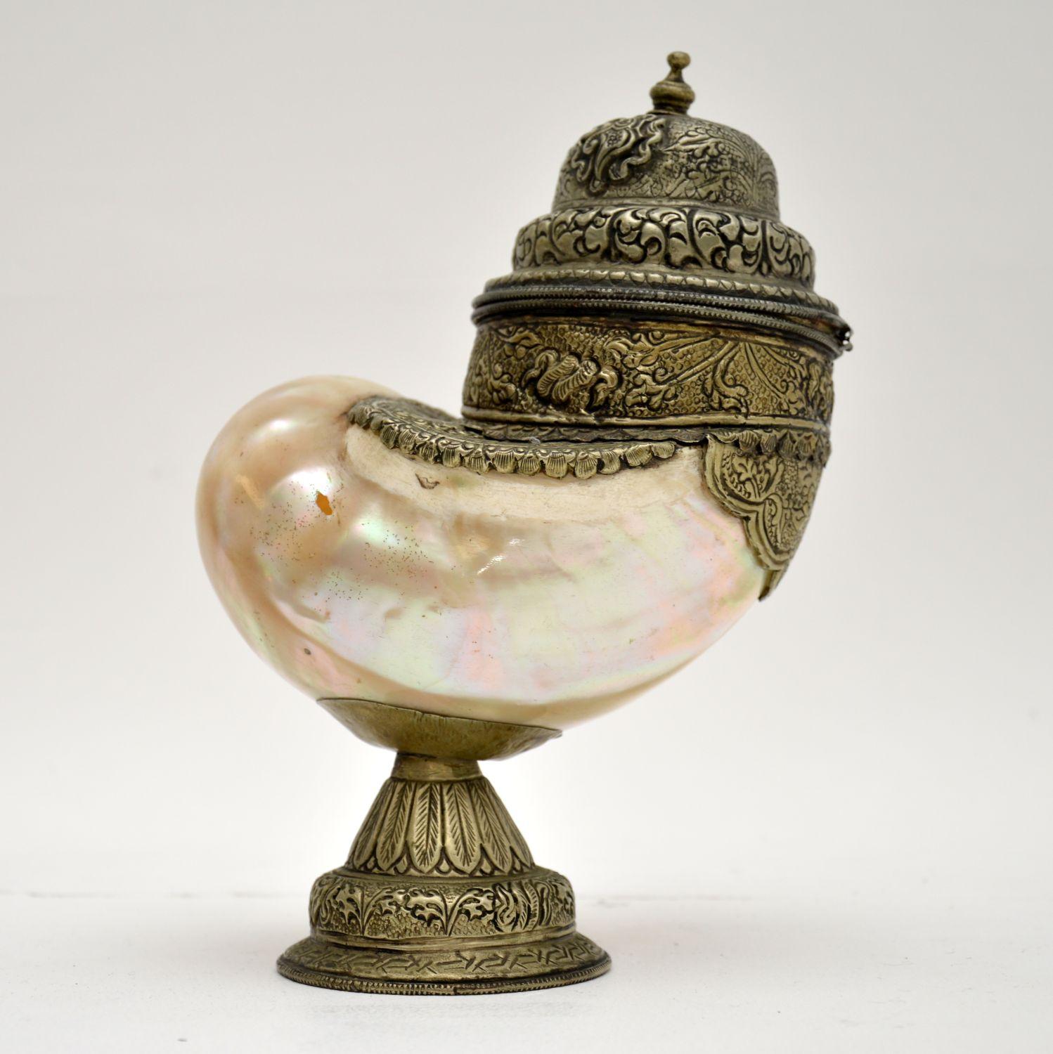 A rare and beautiful ornament, this is an antique Nautilus shell cup with lid, mounted with silver. It was likely made in India, and dates from the late 19th century.
It is beautifully made with fine details, the shell has stunning iridescent