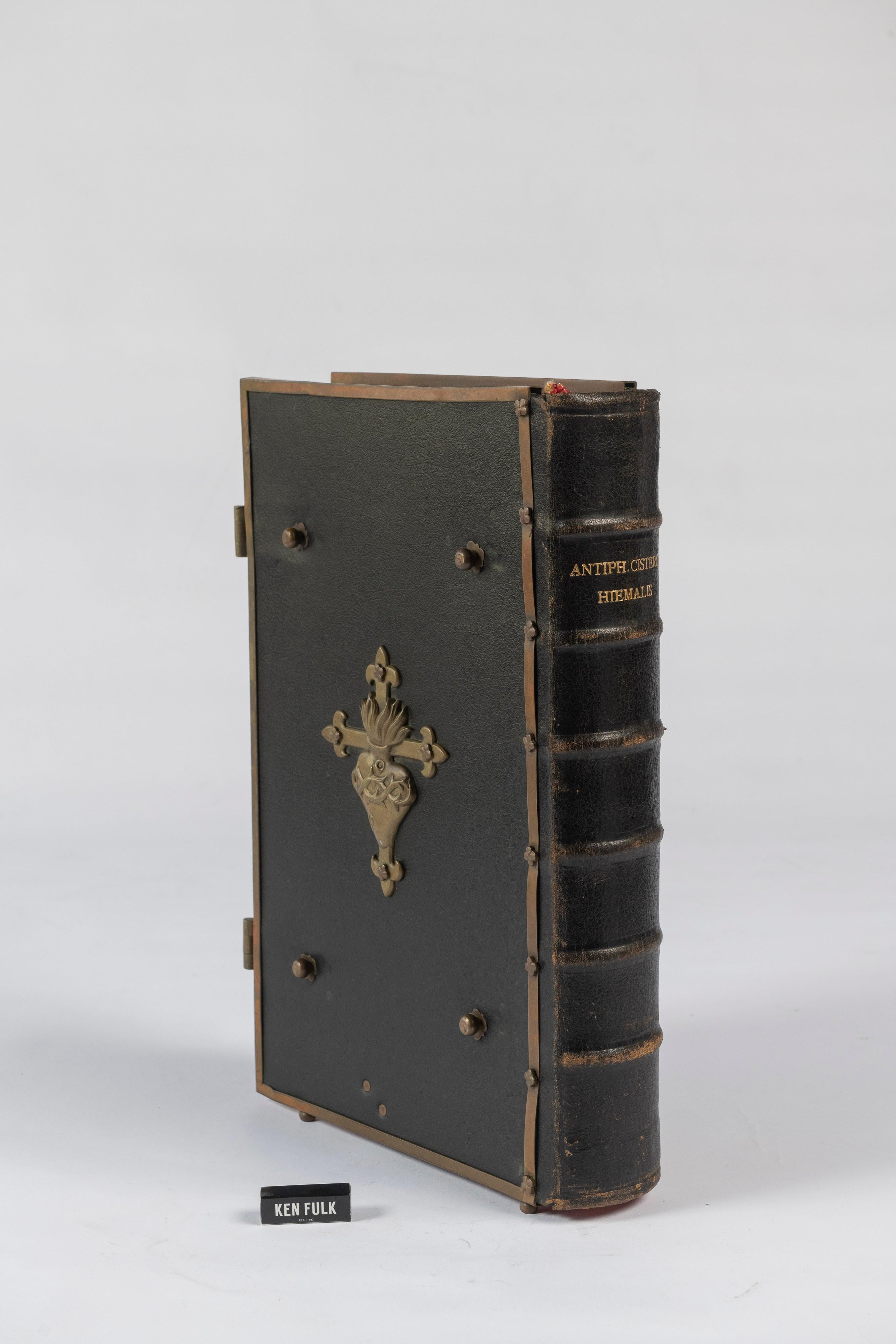 Published by Westmalle of Belgium in 1903, this elaborate hardcover choir book is black morocco over board with metal fore edge clamps, large metal holy cross centerpiece to the upper board, covers framed in metal. The book is printed in black and