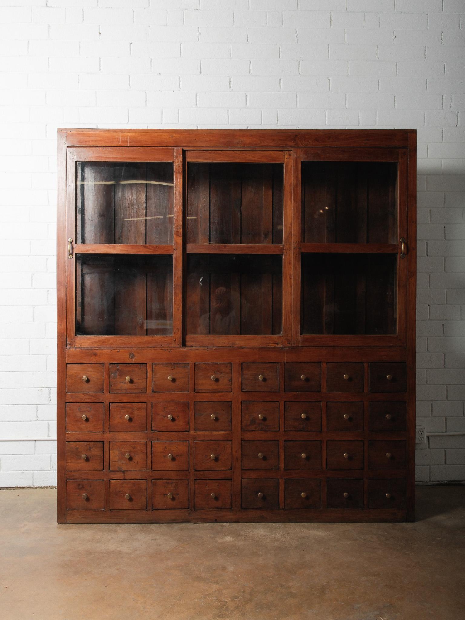 Ein wunderschöner antiker Apothekerschrank mit 32 kleinen Schubladen unter drei Schiebetüren mit Glasfront. Das aus massivem Teakholz gefertigte Möbelstück vereint Funktionalität und zeitloses Design. Die Schubladen - alle mit Messingknöpfen