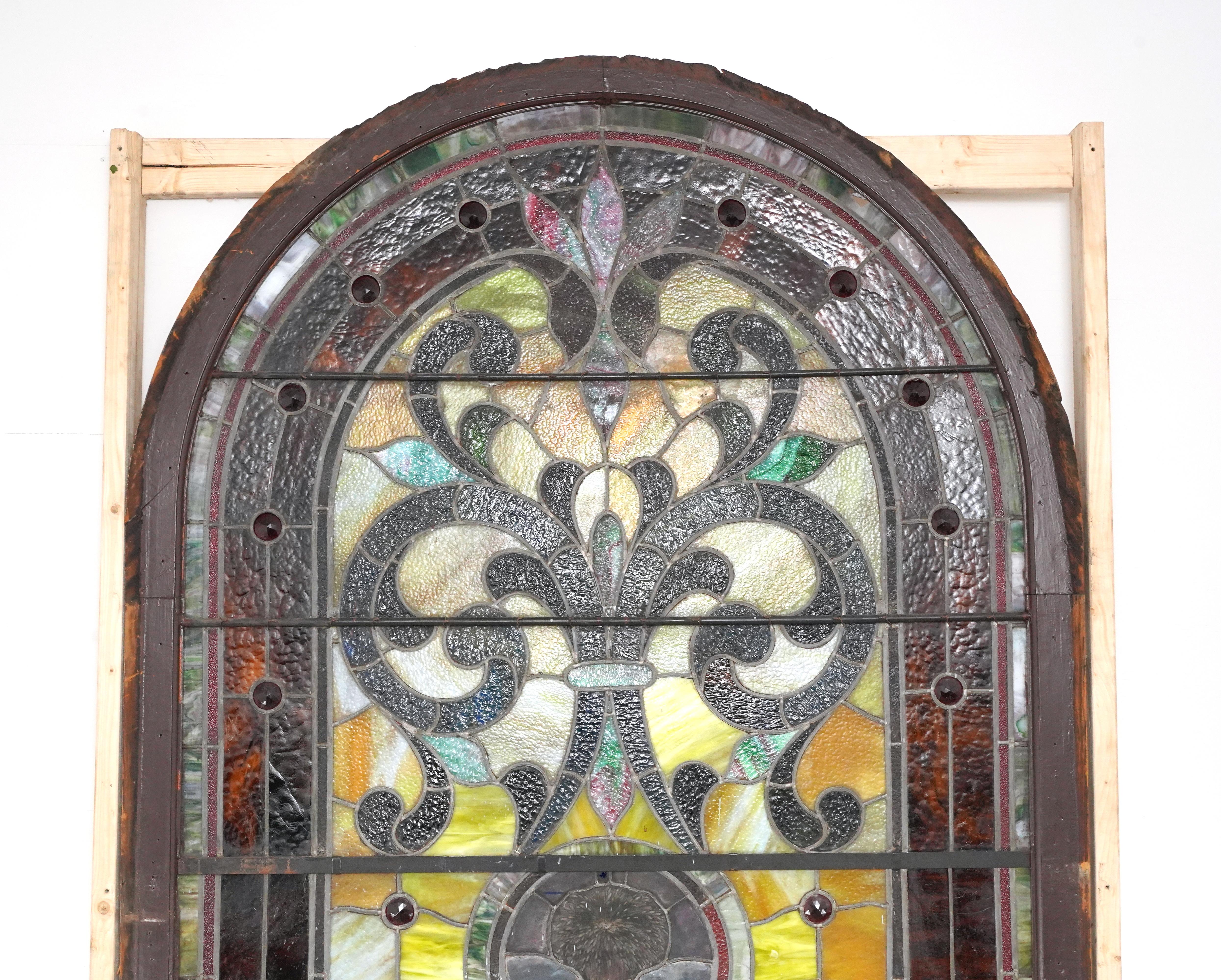 Vitrail d'église en forme d'arche, récupéré, composé de panneaux de verre texturés multicolores, disposés comme des vignes, des feuillages et des feuilles en spirale, avec des accents de bijoux en relief. Le cadre est peint en brun d'un côté et en