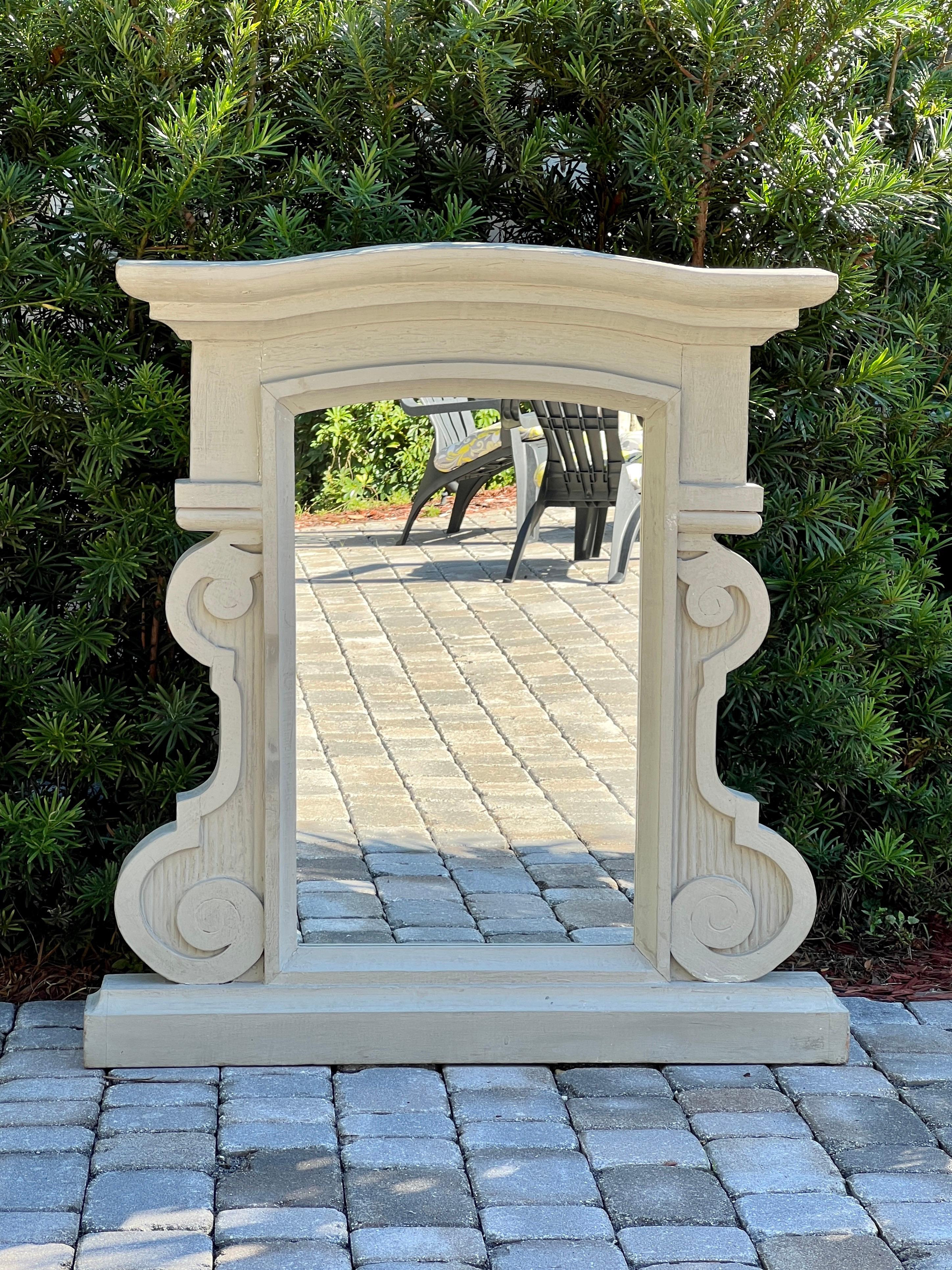 Antique mirror with hand carved dormer window frame in solid wood. The heavyset architectural frame features large chiseled scrolls with fluted details, a curved pediment top, and a sill or ledge base. Hand-painted in a grey whitewash paint with a
