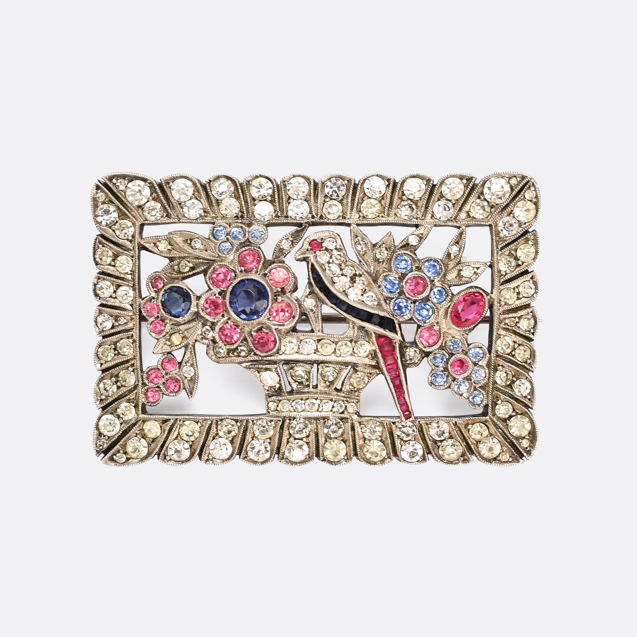 A spectacular Art Deco brooch featuring a giardinetti scene, complete with bird perched on the flower basket, within a picture frame border. It's fully set with lively paste gemstones in white, pink, royal blue, and pastel pink and blue. The