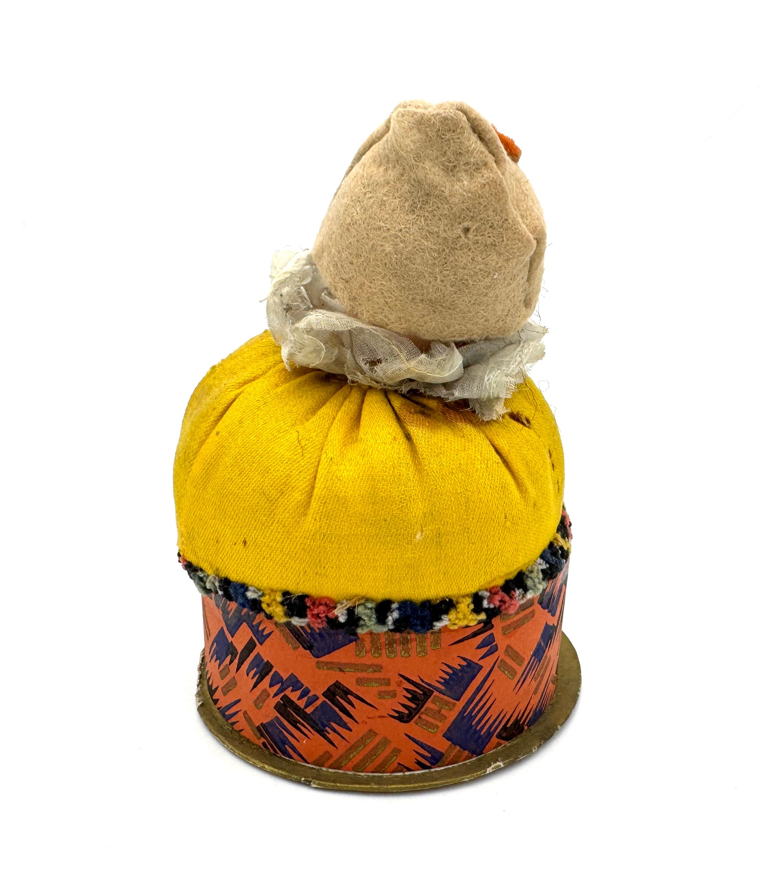 This compact is designed as a pierrette and was hand crafted in 1920 ca. The circular lid of the compact is made from card that is decorated with hand printed Art Deco ornaments. The pierrette wears a beige felt tricorn hat decorated with an oval