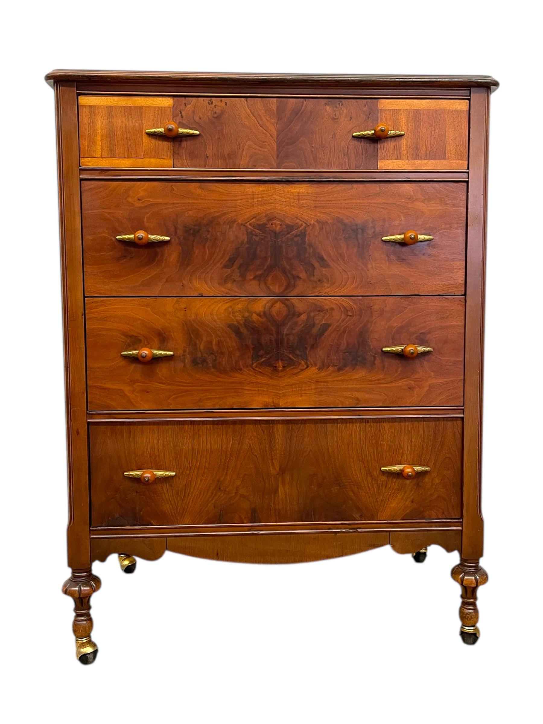 Un comò antico di ottima fattura che mostra le prime influenze dell'Art Déco, con quattro spaziosi cassetti arricchiti dalle caratteristiche maniglie in bachelite e ottone. Le ricche venature del legno e i motivi simmetrici dell'impiallacciatura