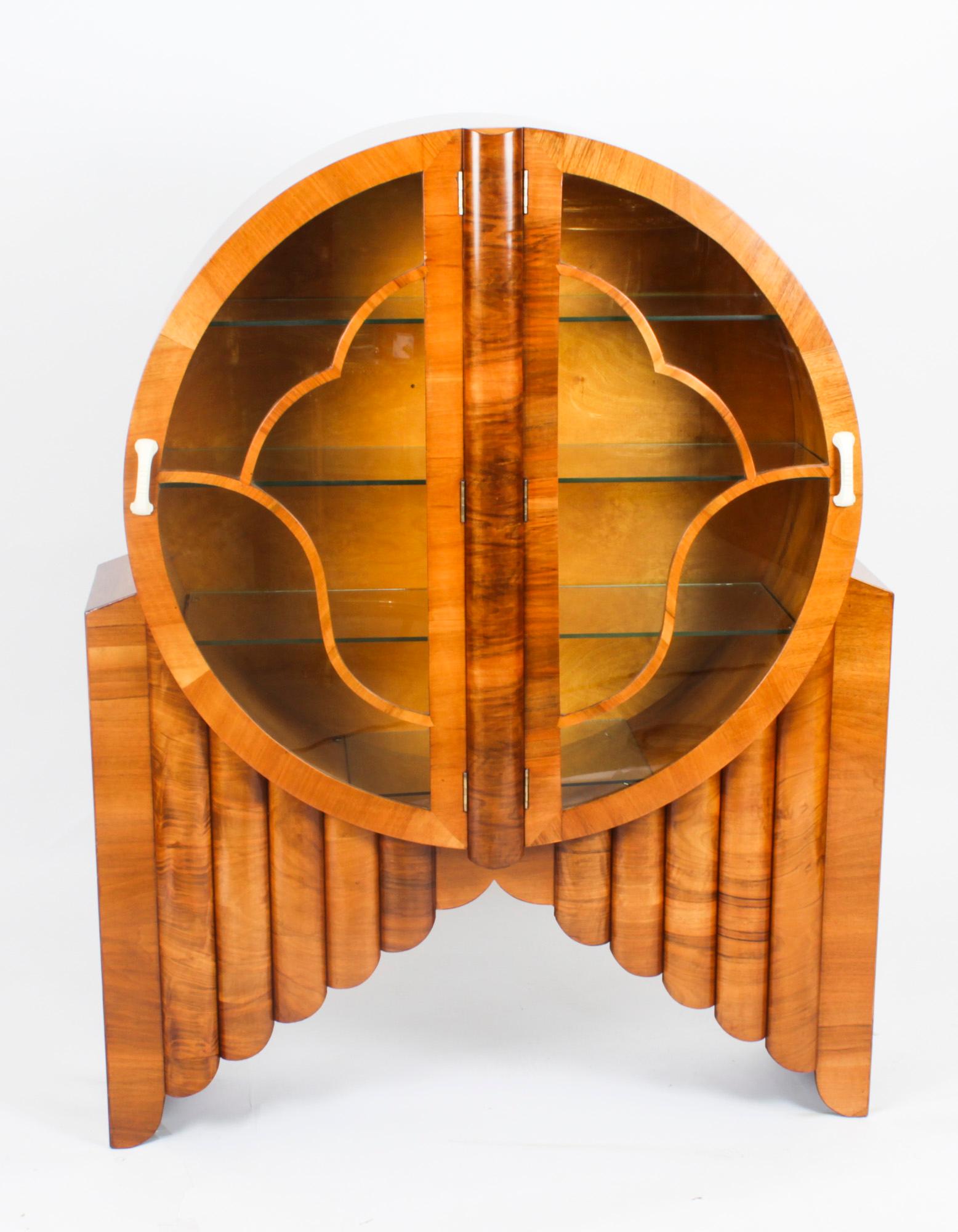 This is a lovely, antique Art Deco figured walnut 'Rocket' display cabinet, circa 1920.
 
The circular cabinet with hinged panelled glazed doors to the centre which enclose four glass shelves, with fretwork beadwork.

Raised on a fluted