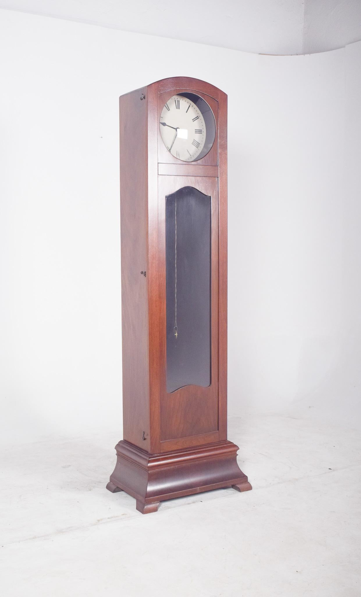 Tall case clock, Deco. The circular dial with classic Roman numerals is set into the top of the cabinet, slightly arched, giving it a touch of softness within the sobriety of the whole. The case is marked by a central glazing that allows a glimpse