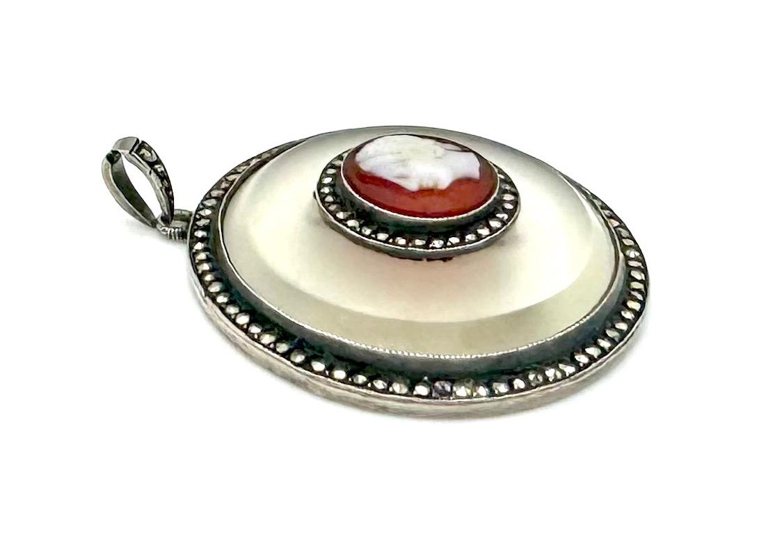 Ce pendentif a été créé vers 1920. Il est en argent sterling. Dans un cadre extérieur serti de marcassites et décoré sur le bord de mille griffes, on voit une plaque ronde biseautée en verre dépoli montée dans un cadre argenté avec un décor de mille