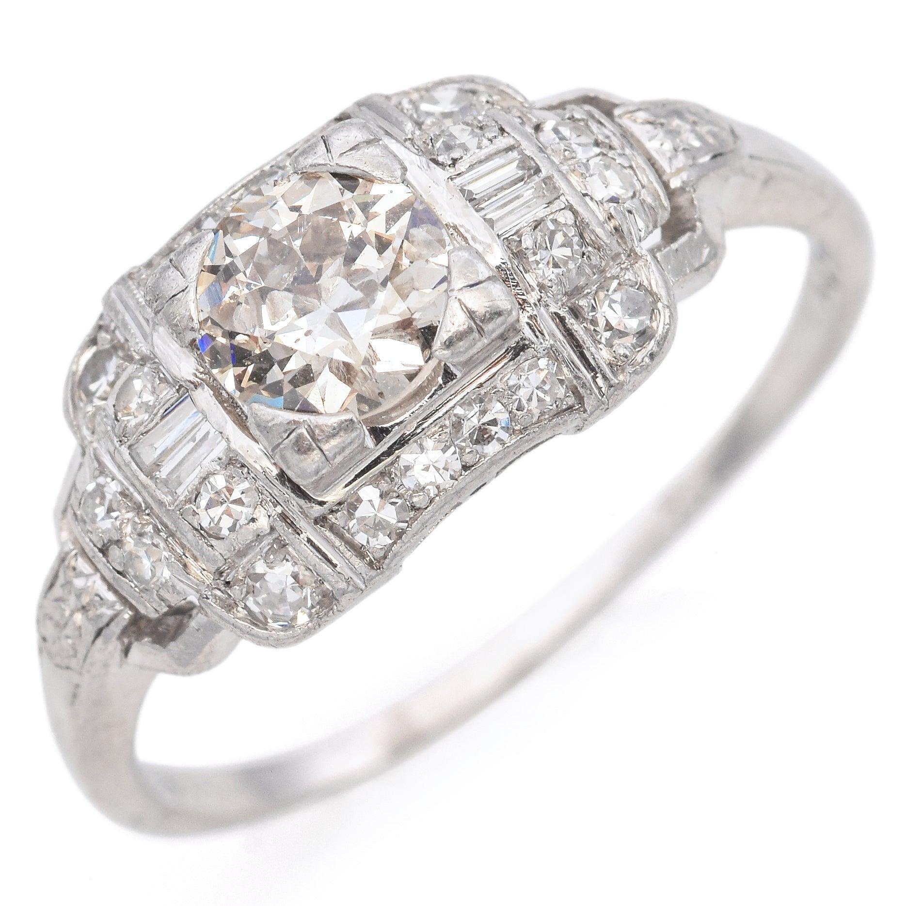 Un anello Art Deco audace ed elegante realizzato in platino, con un diamante centrale scintillante circondato da una disposizione strutturata di pietre rotonde e baguette. Il design geometrico e la raffinata lavorazione del metallo riflettono il