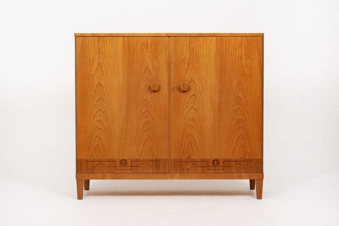 Este precioso armario o aparador sueco antiguo de estilo Art Déco de Carl Malmsten data de 1940. Este mueble está fabricado por expertos en madera maciza y chapa de madera de olmo, y presenta una hermosa banda decorativa de incrustaciones