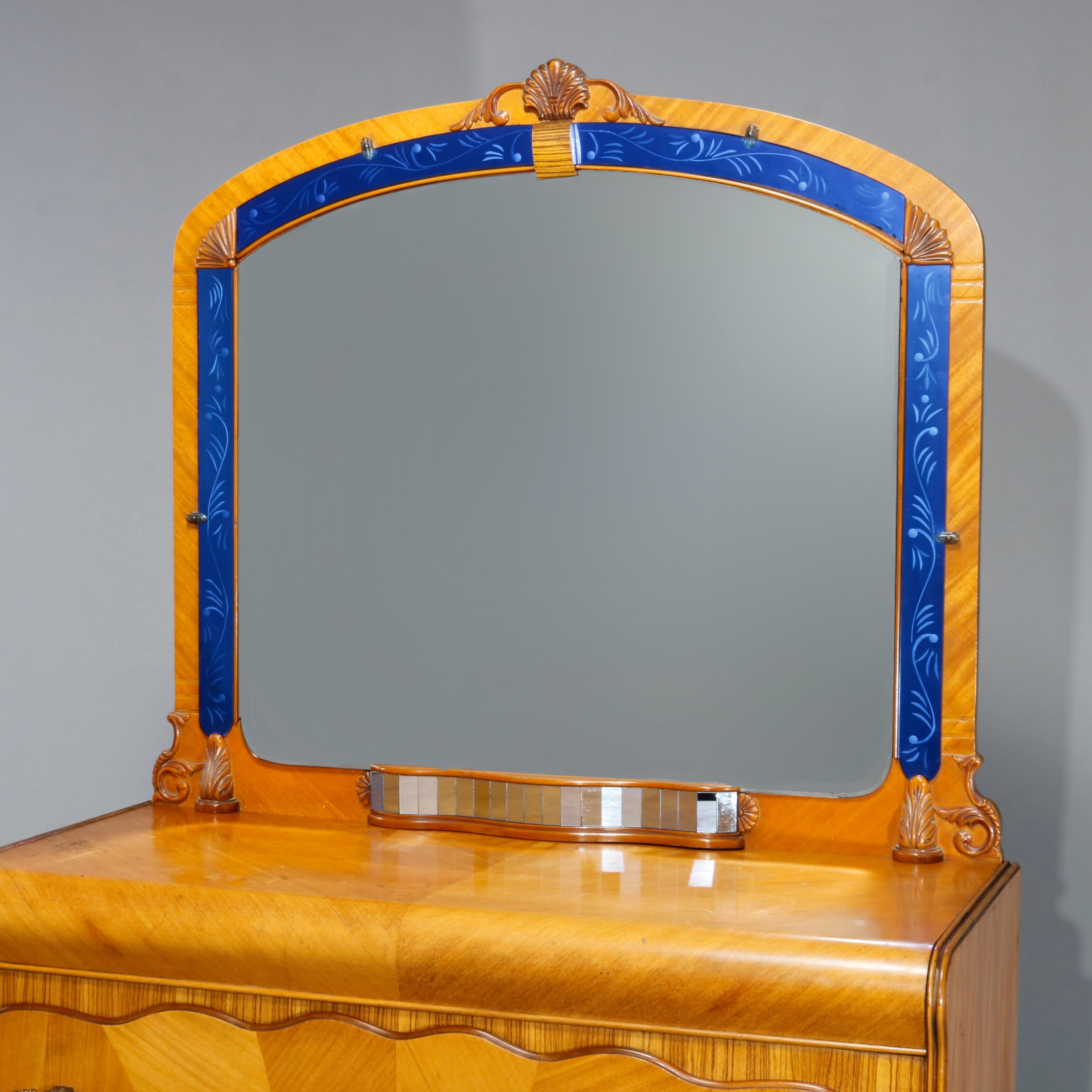 An antique Art Deco waterfall dresser offers mahogany construction with shaped mirror having shell carved crest and foliate etched blue glass bordering surmounting chest of drawers with satinwood sunburst inlay across three long drawers, stylized
