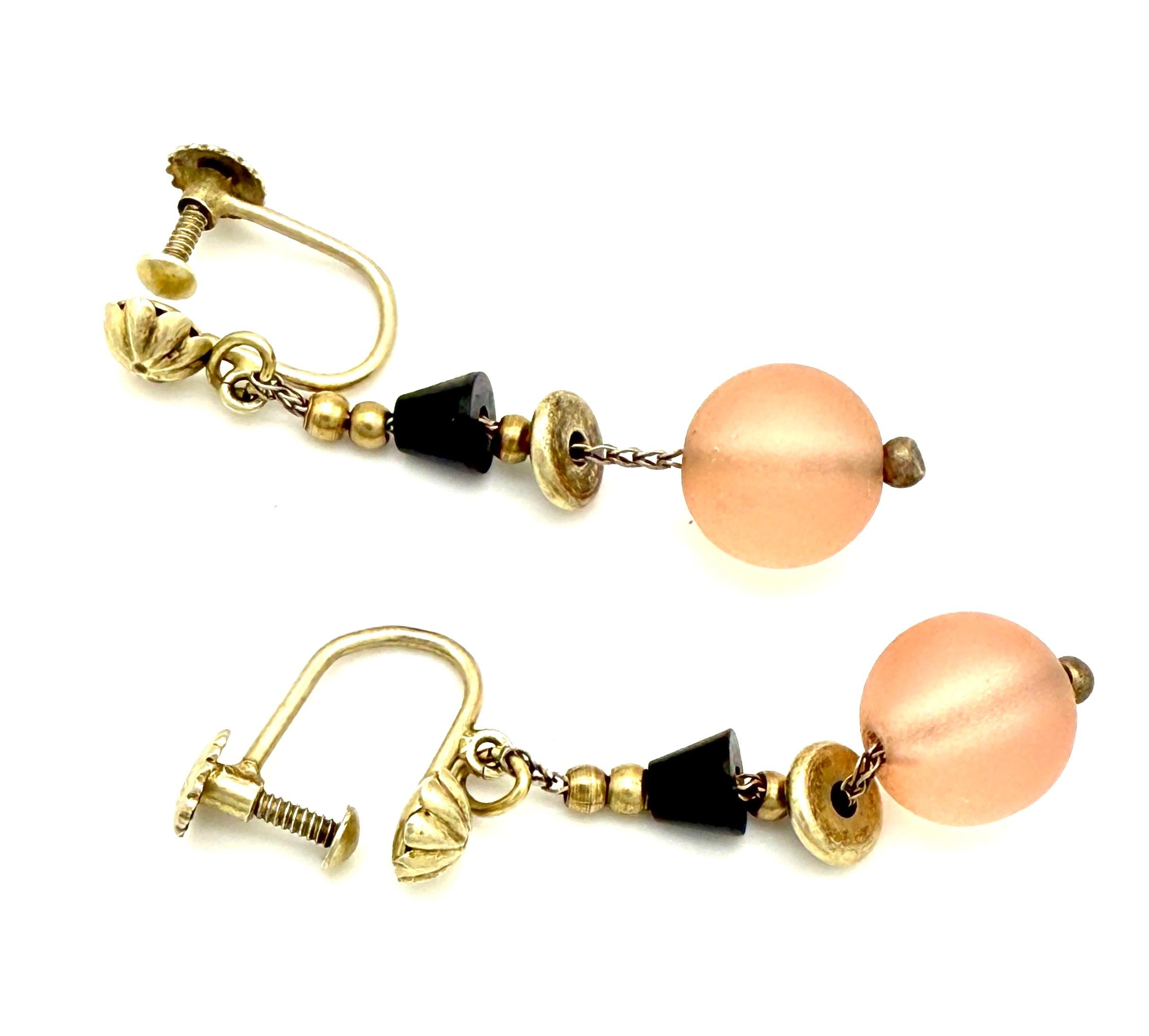 Questi insoliti orecchini pendenti in argento Art Deco sono stati realizzati nel 1920 circa.
Due perle di vetro smerigliato di colore rosa WMF Ikora sono infilate in una catena a coda di volpe in argento dorato insieme a perle nere a forma di