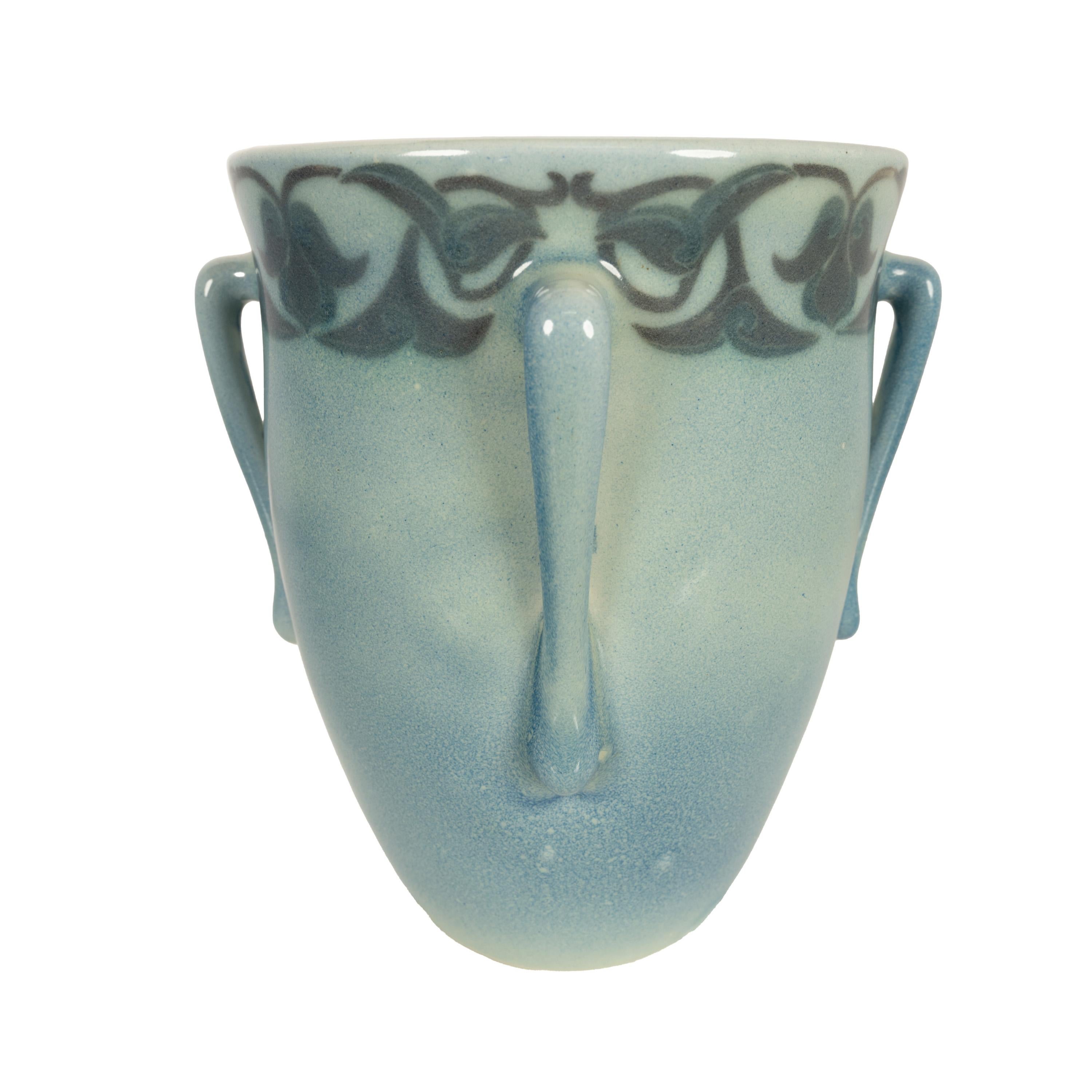 Antico vaso americano Art Nouveau Rookwood a tre manici in vellum, Lorinda Eppli, 1915.
Il vaso blu a forma di campana rovesciata con tre manici allungati, intorno all'ampia bocca del vaso è presente una fascia di decorazione floreale stilizzata blu
