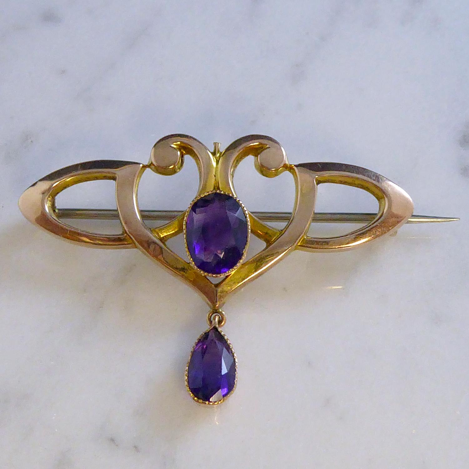 A antique brooch from the Art Nouveau era and dating from the early 1900s.  An oval mixed cut amethyst of dark purple colour is set in a milled grain edge collar mount to the centre of an ornate, open scroll rose gold design.  Suspended from the