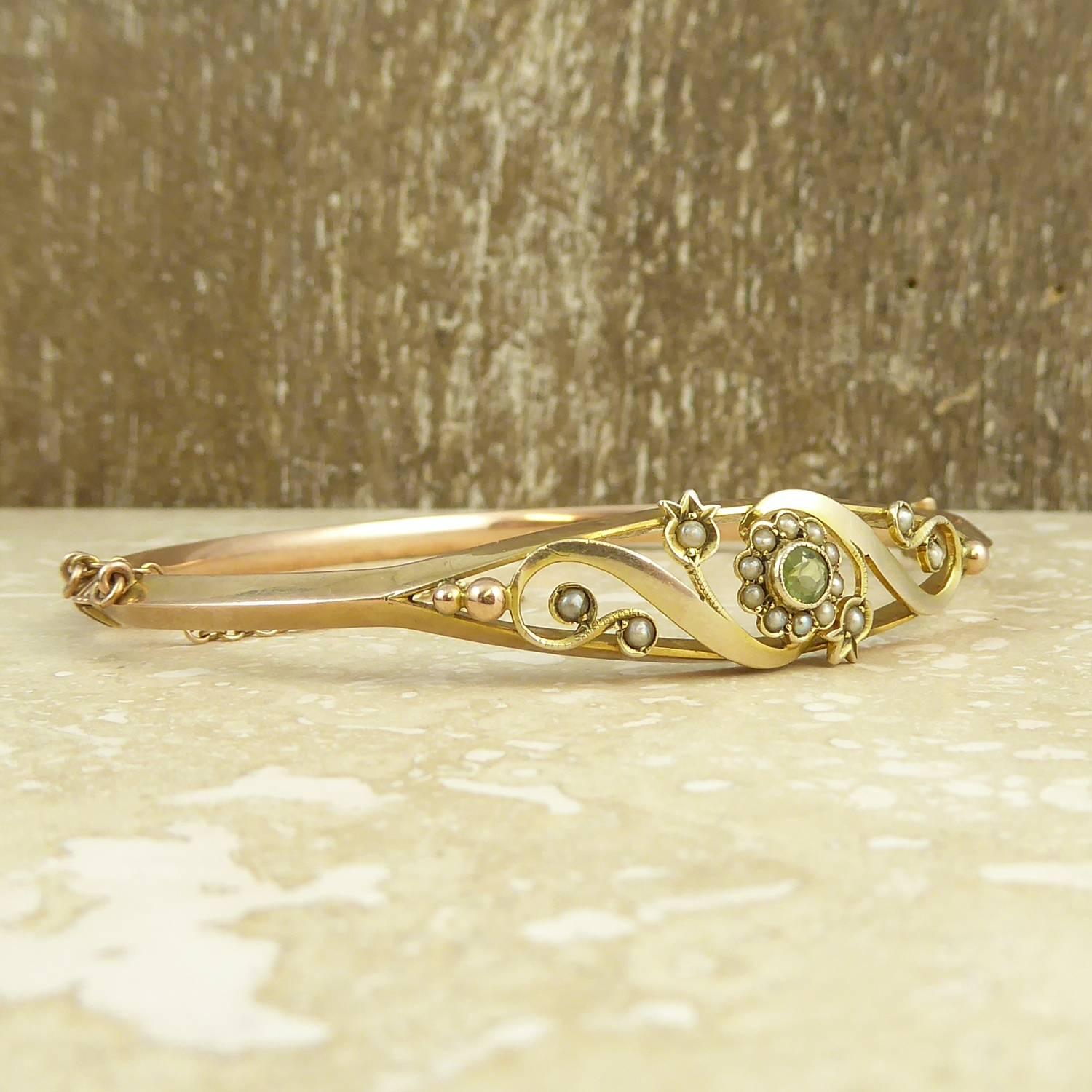 An antique gold bangle from the romantic Art Nouveau era and hallmarked for the year 1911.  The bangle has an open boat-shaped front which is set to the centre with a peridot and pearl flower cluster with pearl leaves and surrounded by scroll