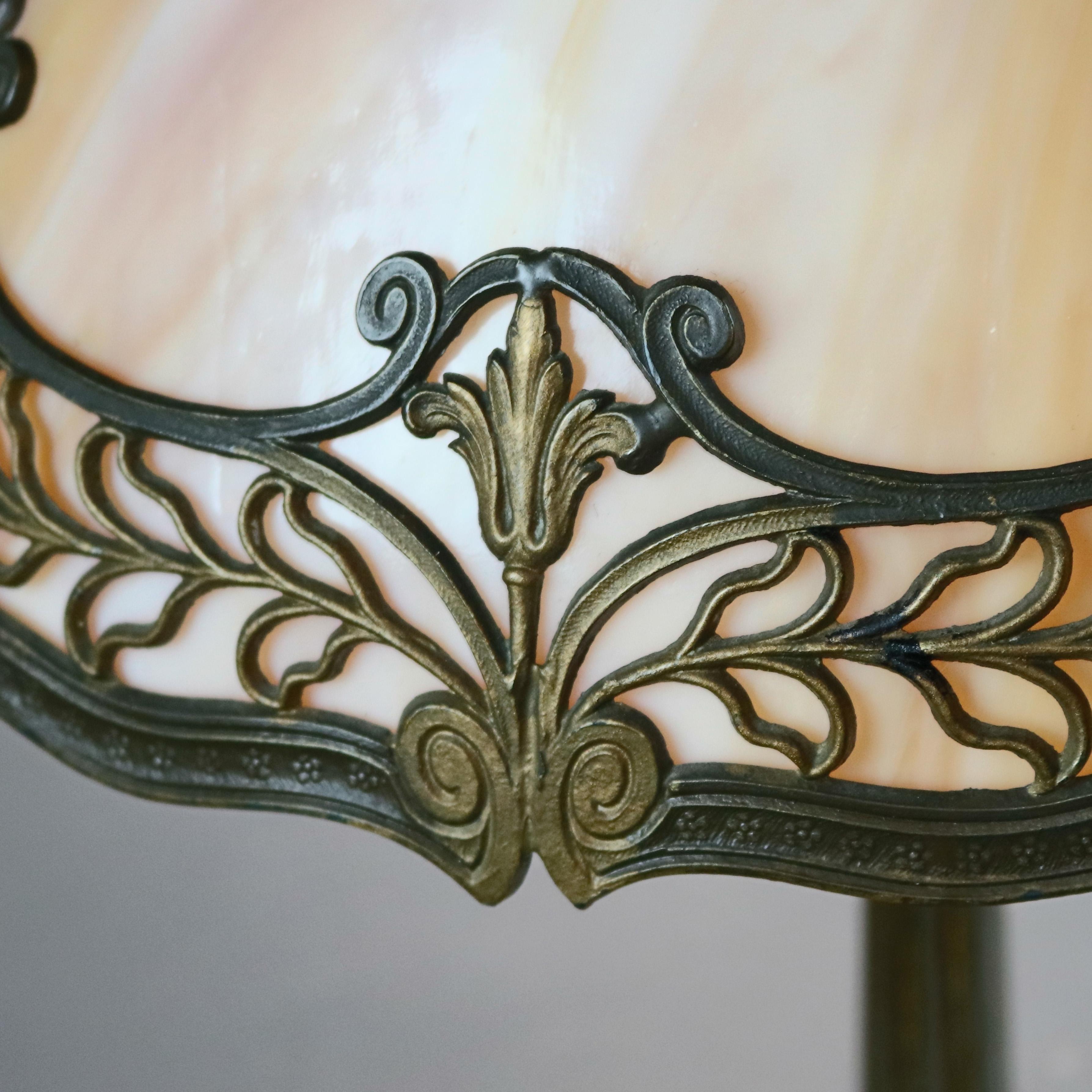 An antique Art Nouveau Bradley & Hubbard school table lamp offers polychromed cast foliate filigree paneled shade housing bent slag glass surmounting cast scroll and foliate form base having two independently controlled light sockets, circa
