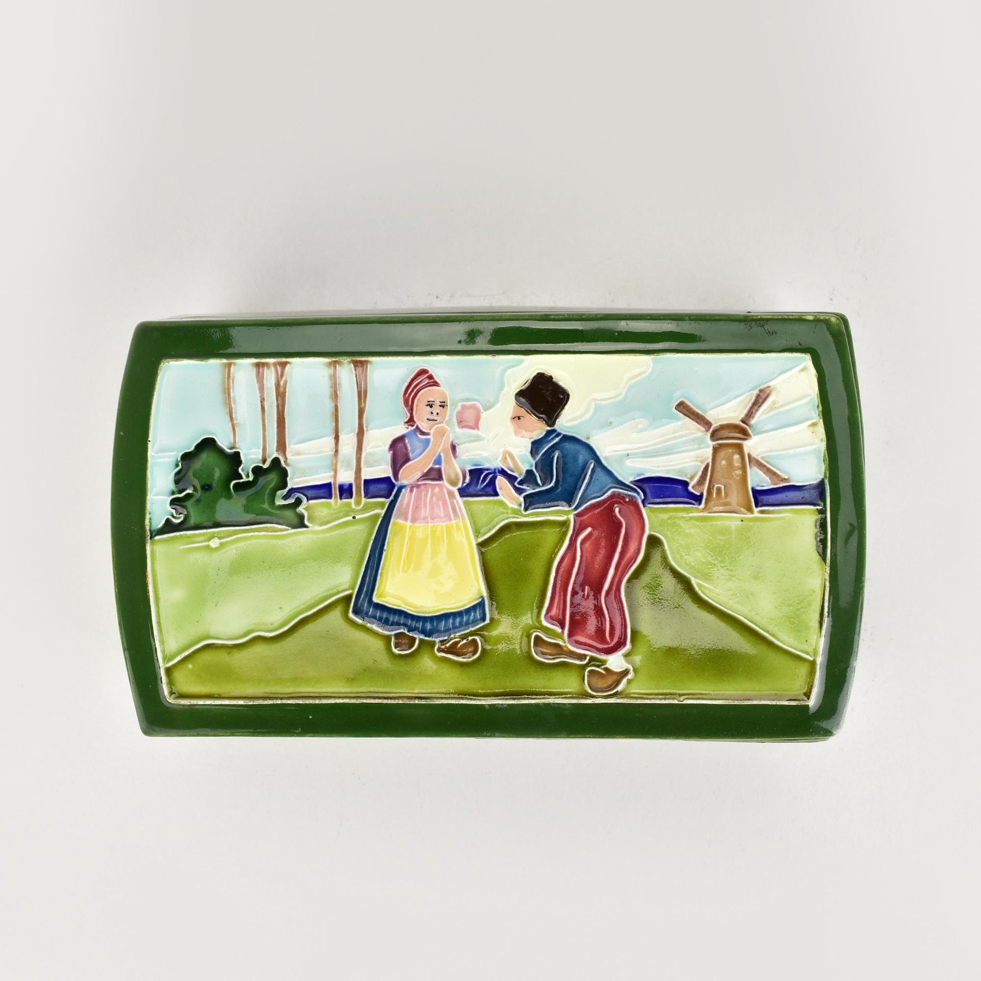 This exquisite antique Art Nouveau ceramic box, designed by Carl Sigmund Luber for Johann von Schwarz, showcases a charming pastoral scene on its lid, featuring a Dutch couple in traditional attire against a picturesque landscape with a windmill.