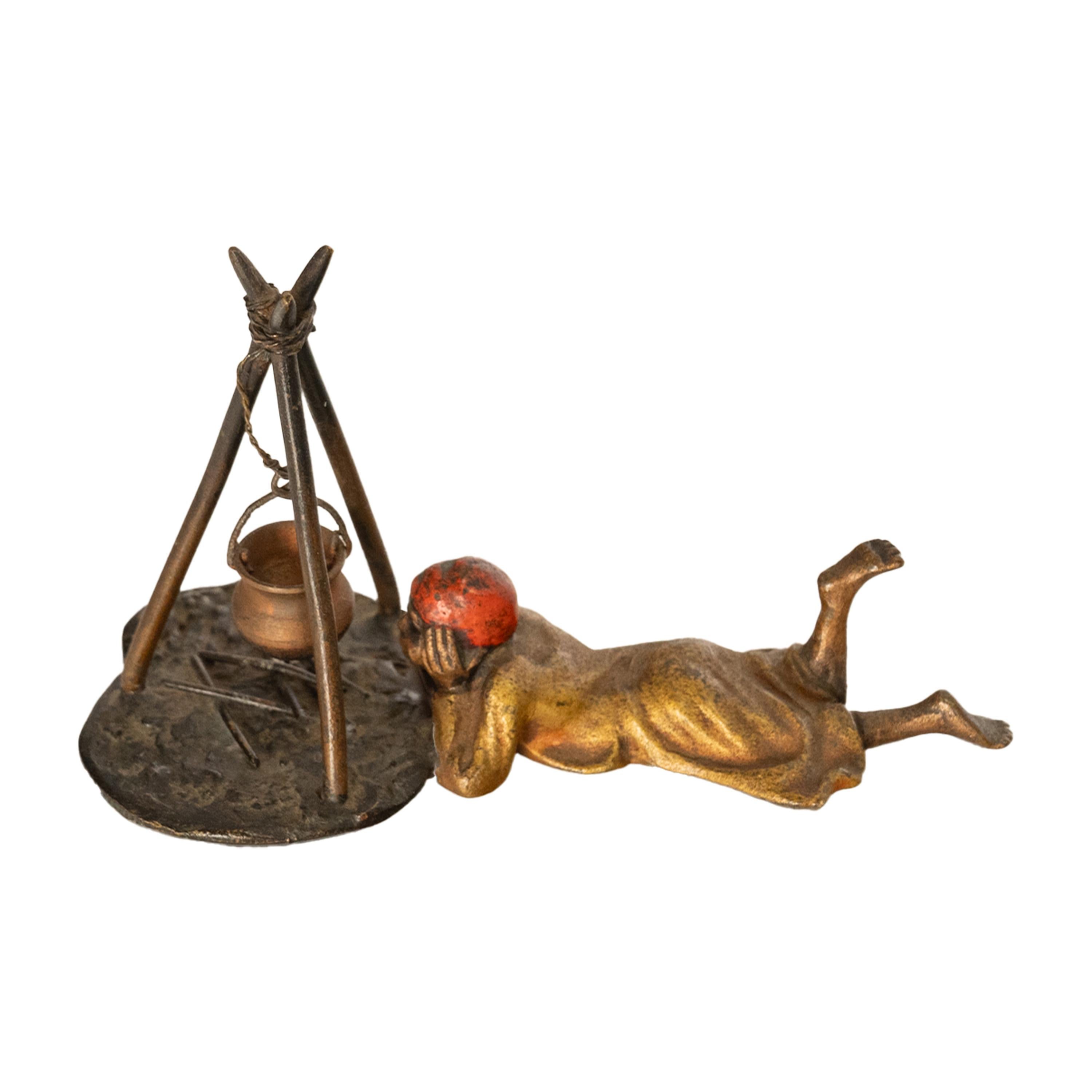 A charming and whimsical antique cold painted bronze in the Orientalist taste, attributed to Franz Bergmann, Austria circa 1910.
The bronze depicts a young Arab boy lying on his front and gazing at his campfire, above is a tripod support with a