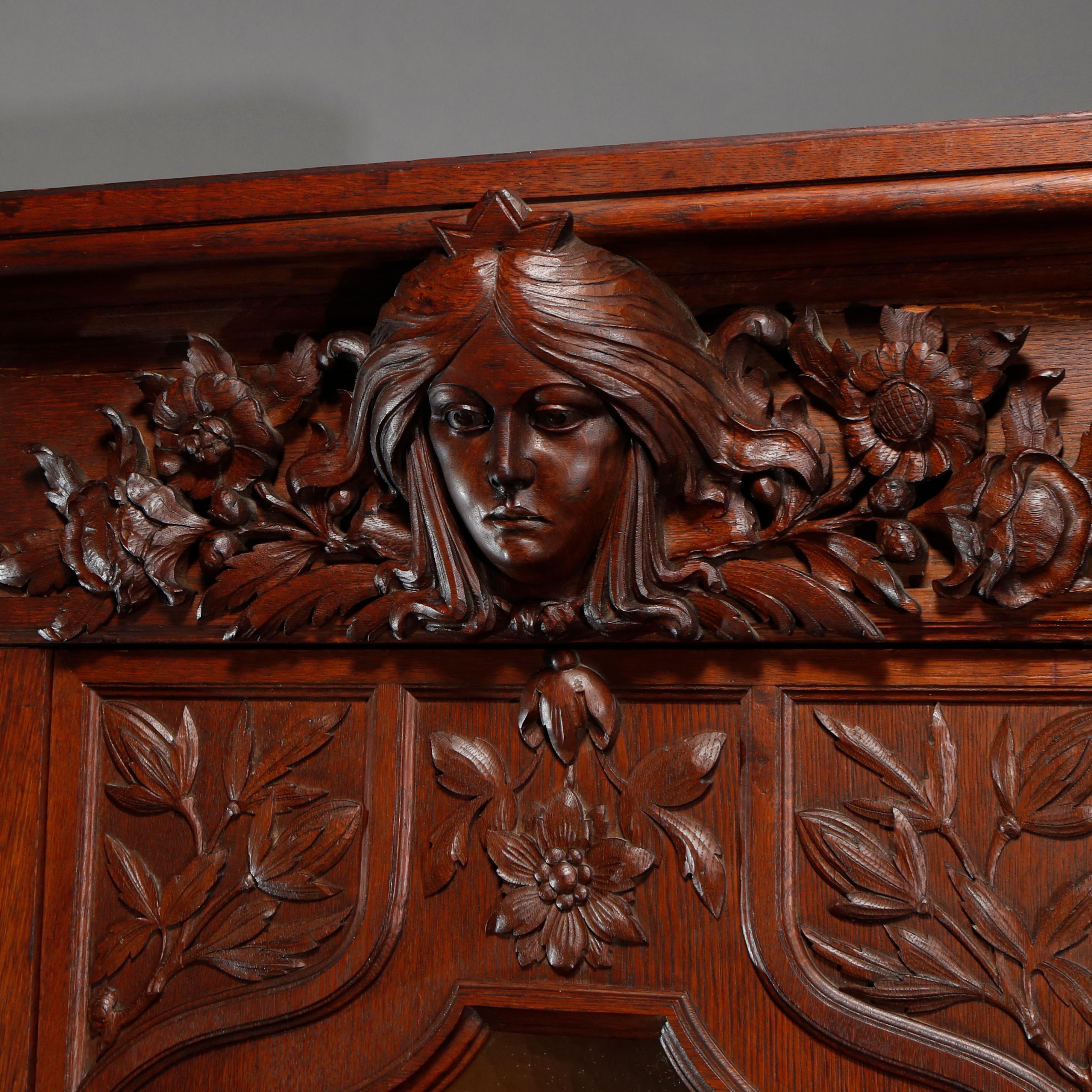 An antique Art Nouveau bookcase offers oak construction with crest having carved Alphonse Mucha style female mask of Sarah Bernhardt with flanking floral elements surmounting case with central cabinet having leaded slag glass door with lily pads