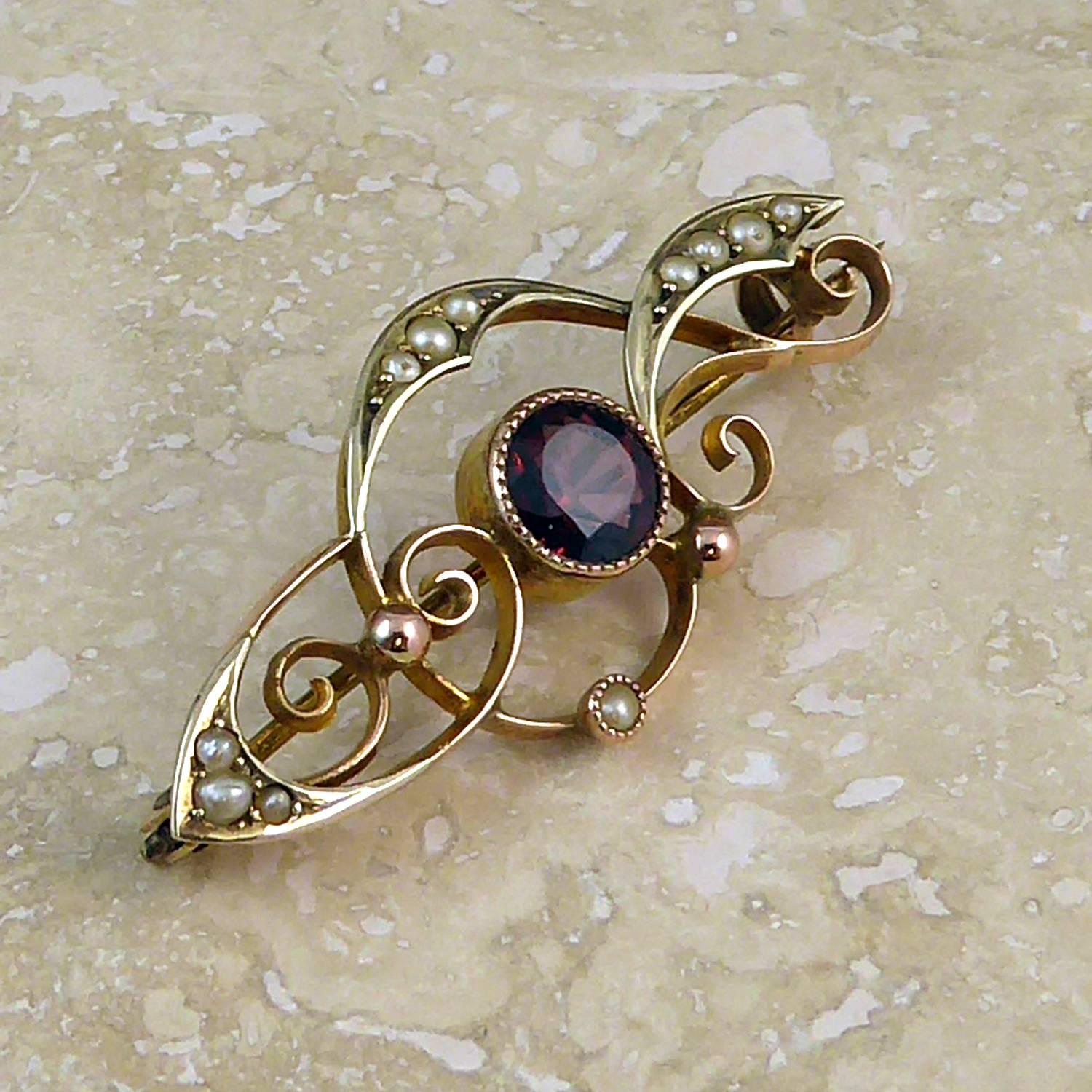 An antique brooch from the early 1900s crafted in the Art Nouveau fashion.  Set to the centre with a round faceted garnet of good purple/red colour, approx. 5.5mm diameter, and mounted in a rose gold collar with decorative edging.  Around the garnet