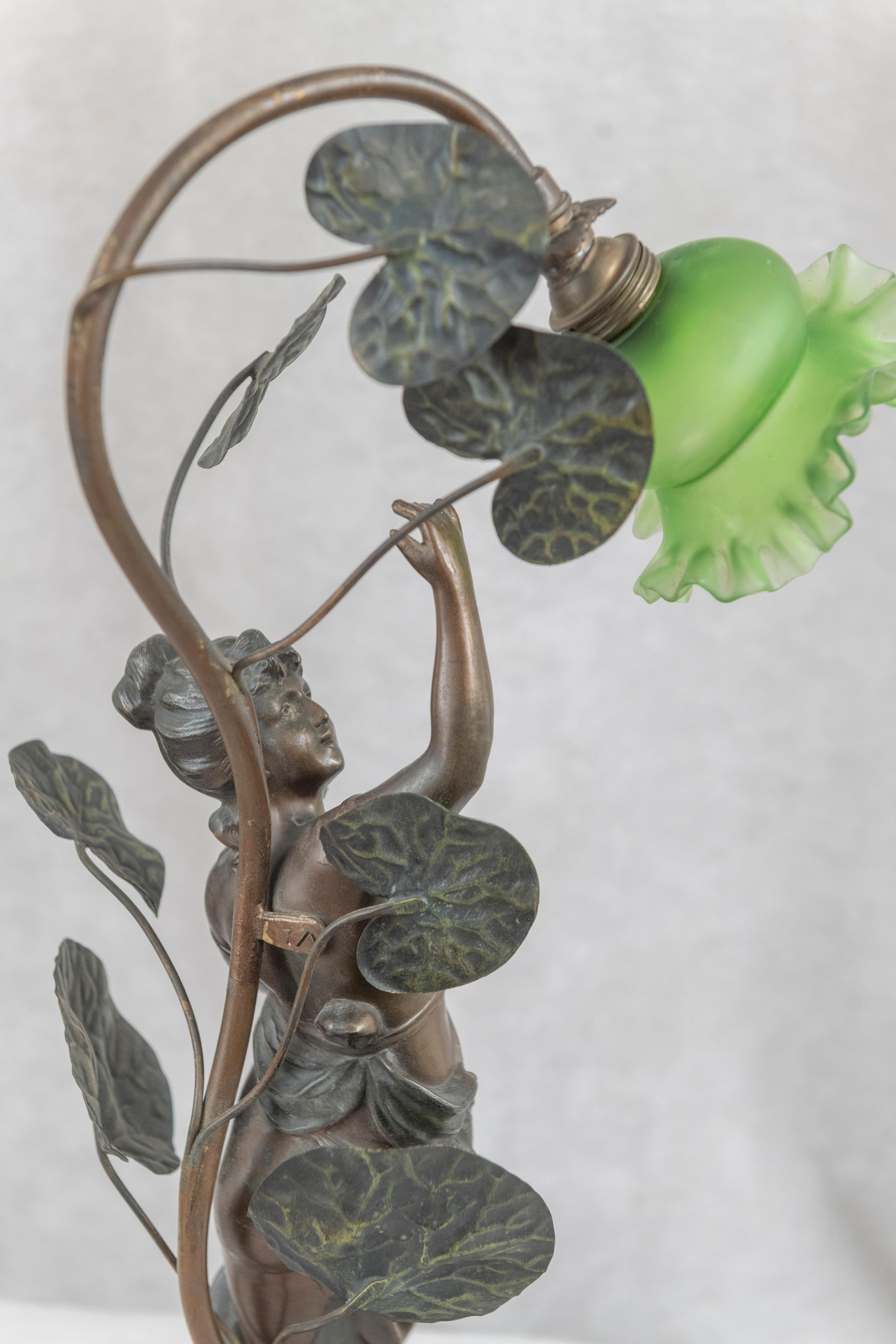 Graceful art nouveau lamp featuring a young lady in nature under a large plant holding the original green glass shade. The sculpture is well modeled and displays a 2 color factory patina. The particularly small green glass shade is hand blown and
