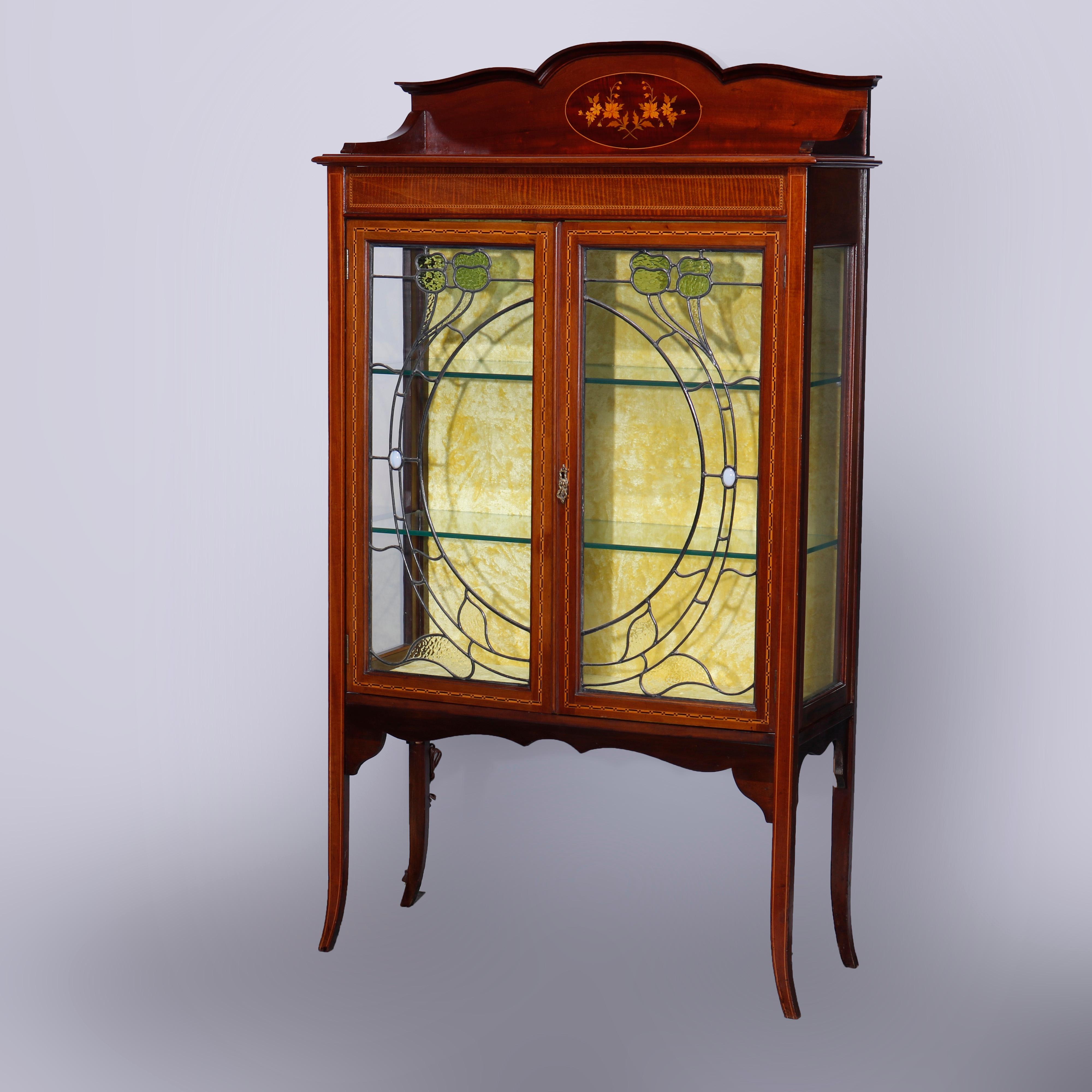 An antique Art Nouveau cabinet offers mahogany construction with shaped backsplash having ebonized trimming and central floral satinwood marquetry inlaid reserve over cabinet having deeply striated frieze surmounting double stained and jeweled glass