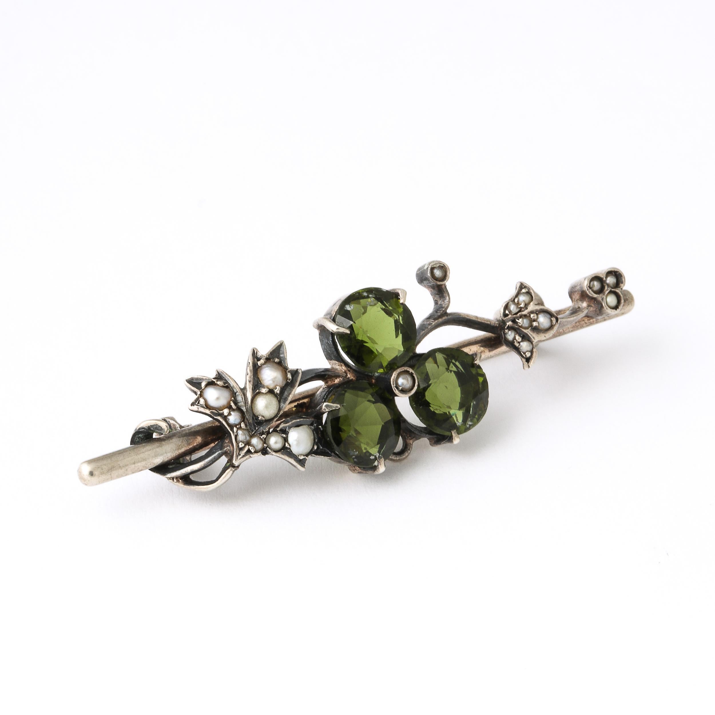 This  Antique Art Nouveau continental silver floral bar pin displays a single flower comprised of 3 round and cut peridots and a central seed pearl with a surround of leaves inlaid throughout with seed pearl accents . It also bears several